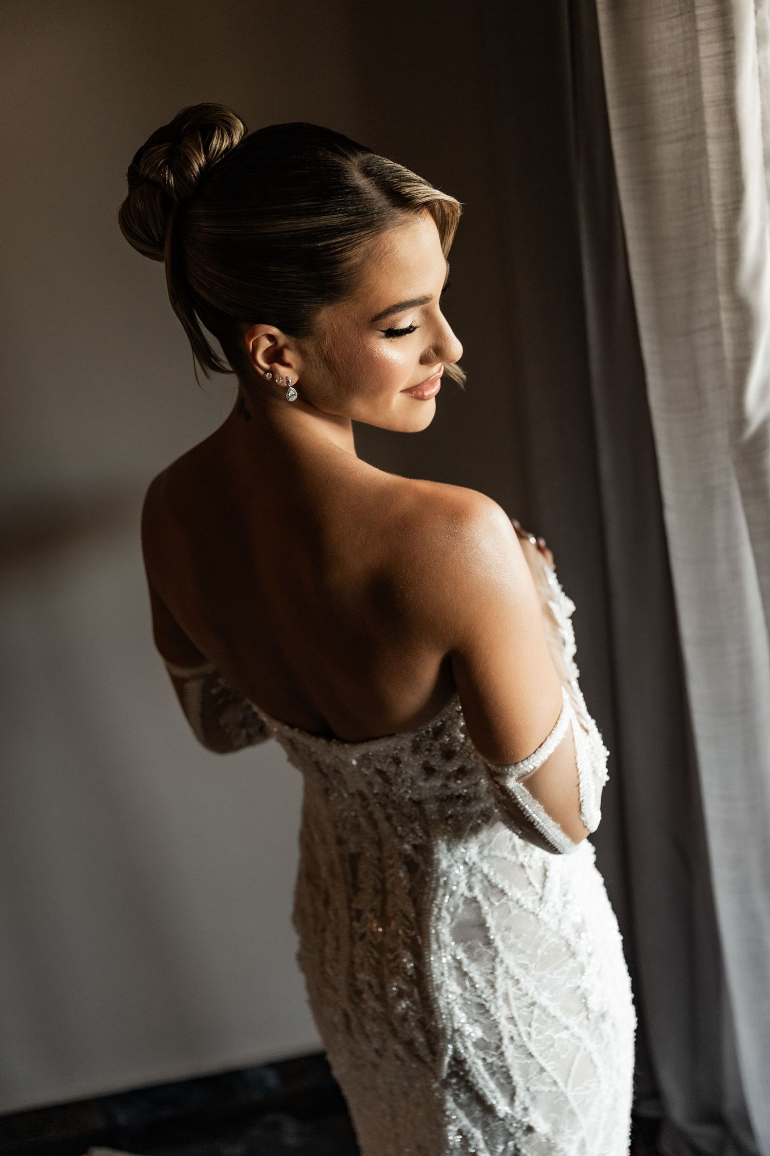 Elegant bride in backless beaded white gown with updo hairstyle posing near window in soft natural light.