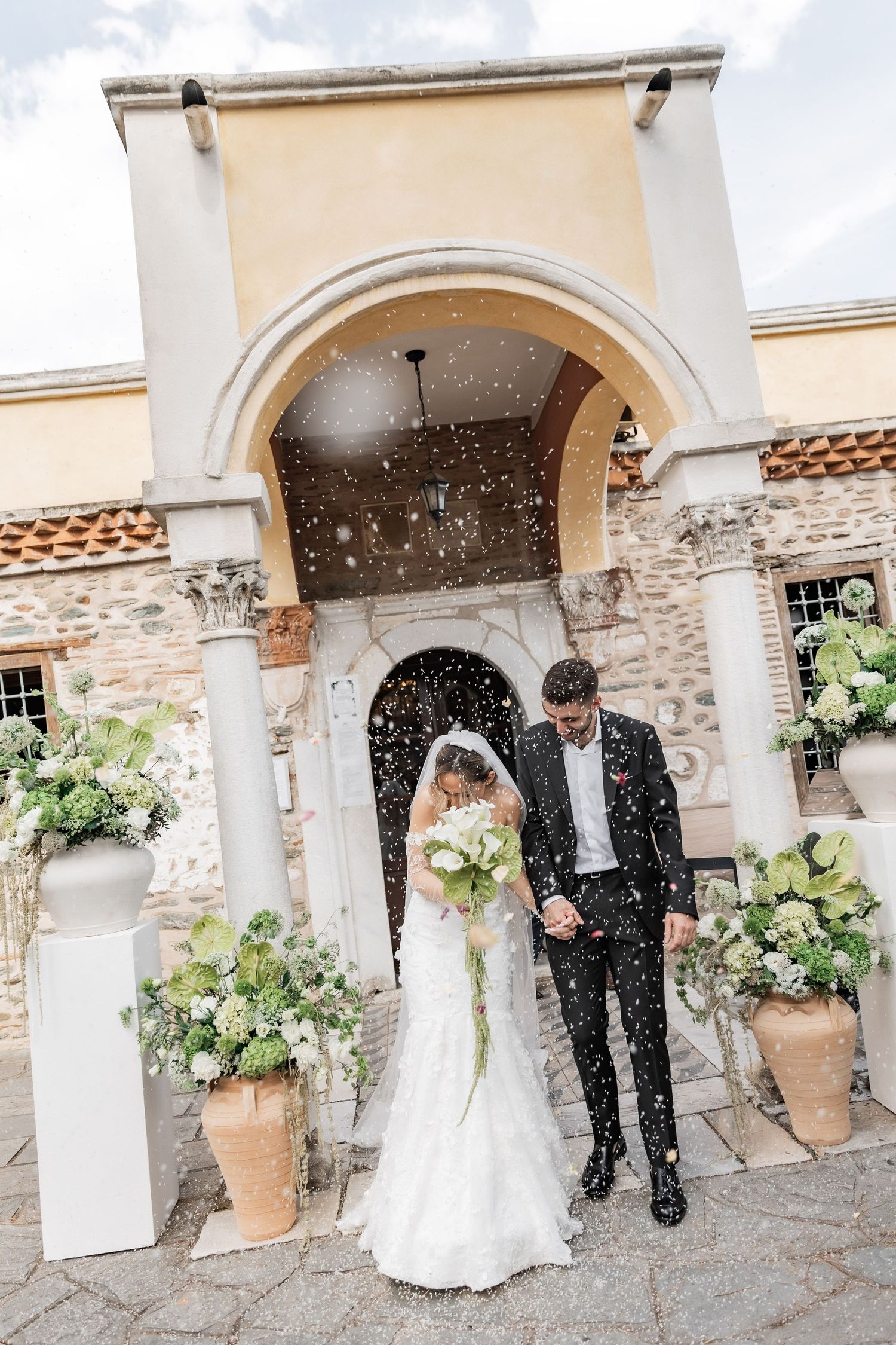 Bride and groom exit a classic church entrance as confetti falls, bride holding a white bouquet.