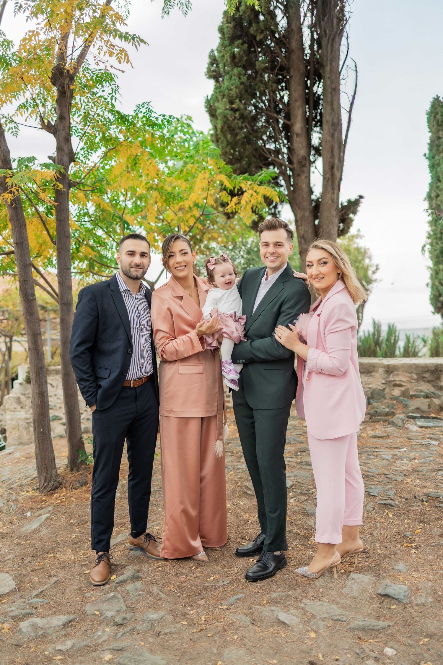 A group of people in formal attire pose together outdoors among autumn trees in coordinated pink and brown outfits.