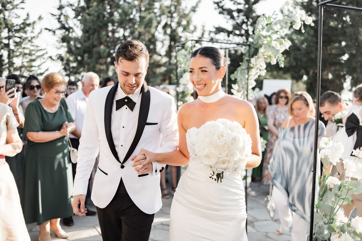 Wedding guests mingle during an outdoor celebration surrounded by pine trees.