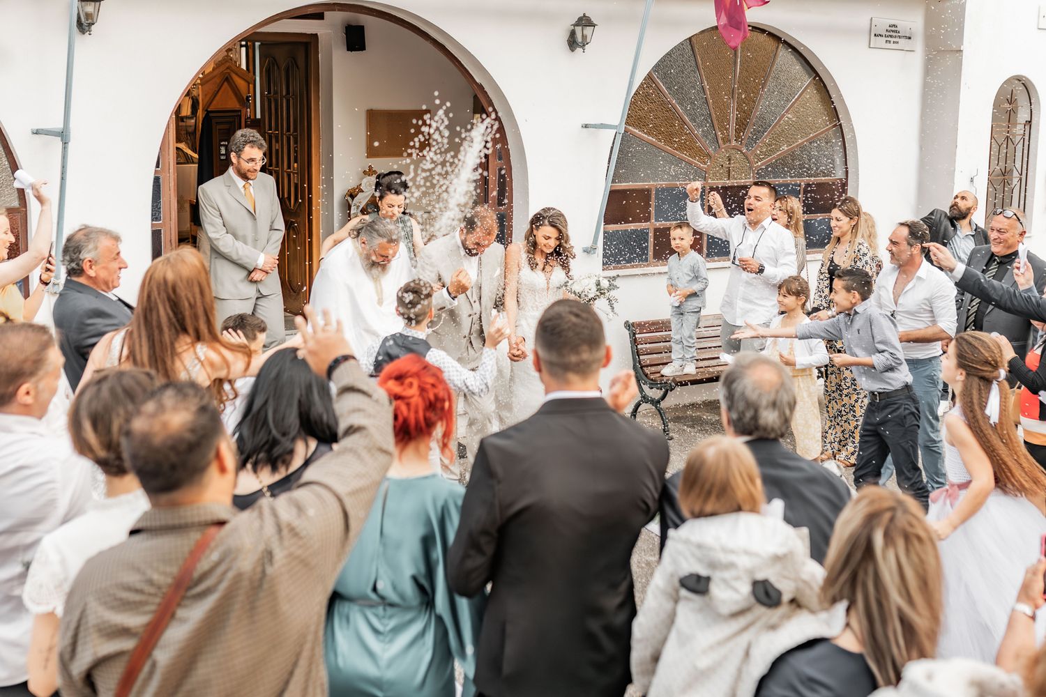 Wedding guests celebrate outside a white building with arched doorways.
