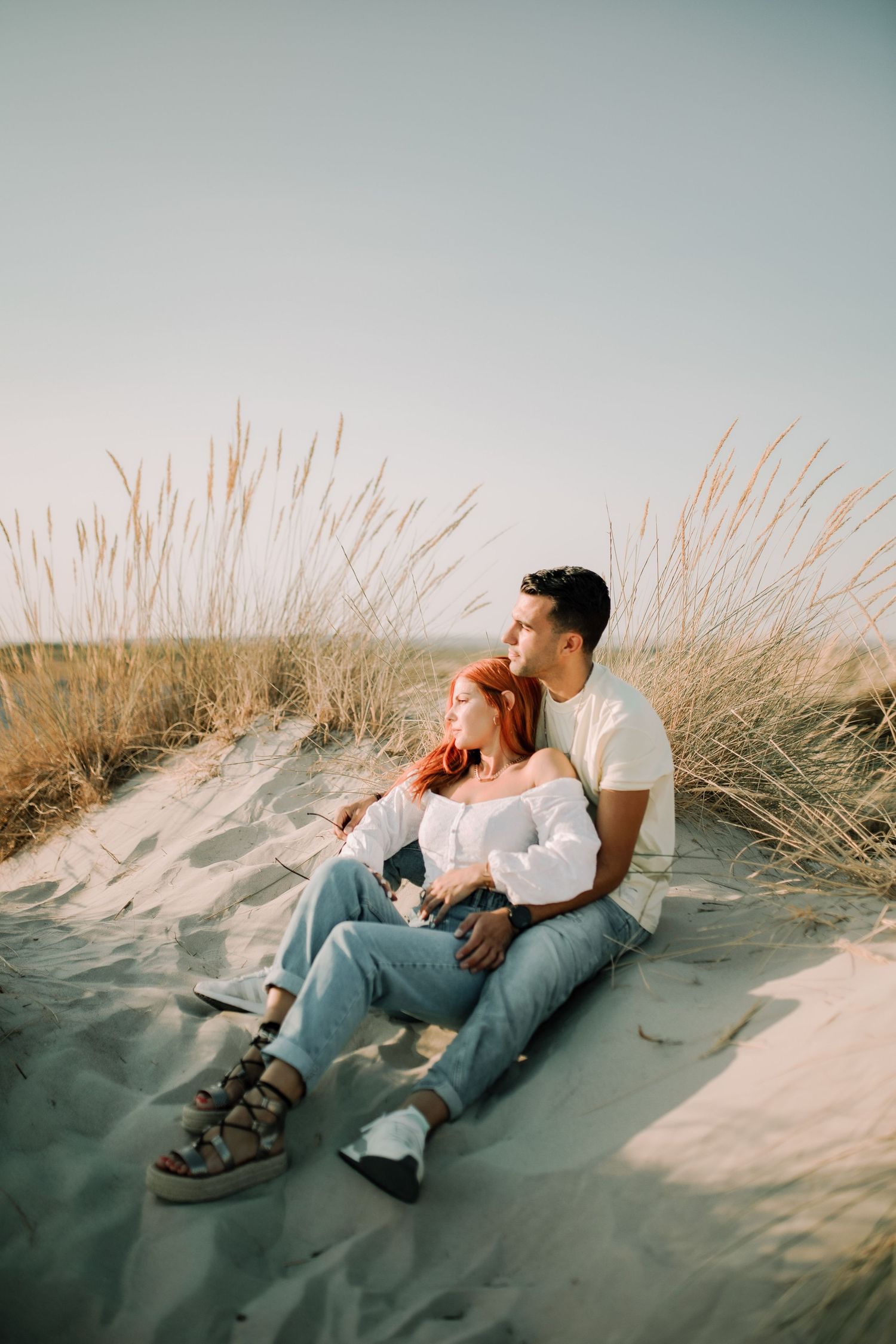 A couple relaxes together on a sandy beach dune surrounded by tall grass during golden hour.