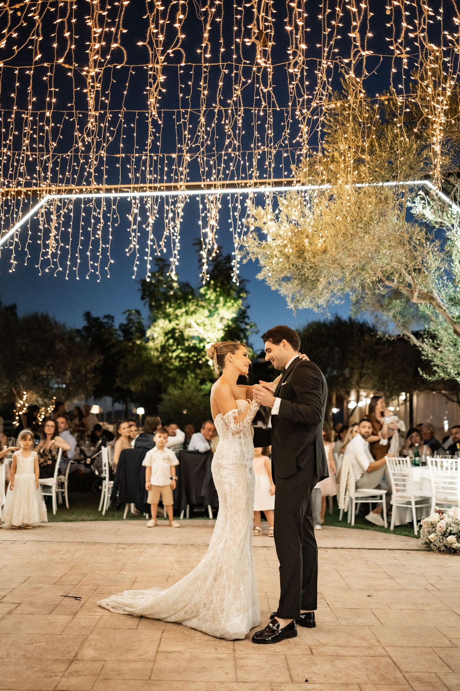 Bride and groom share first dance under twinkling fairy lights at elegant outdoor evening wedding reception.