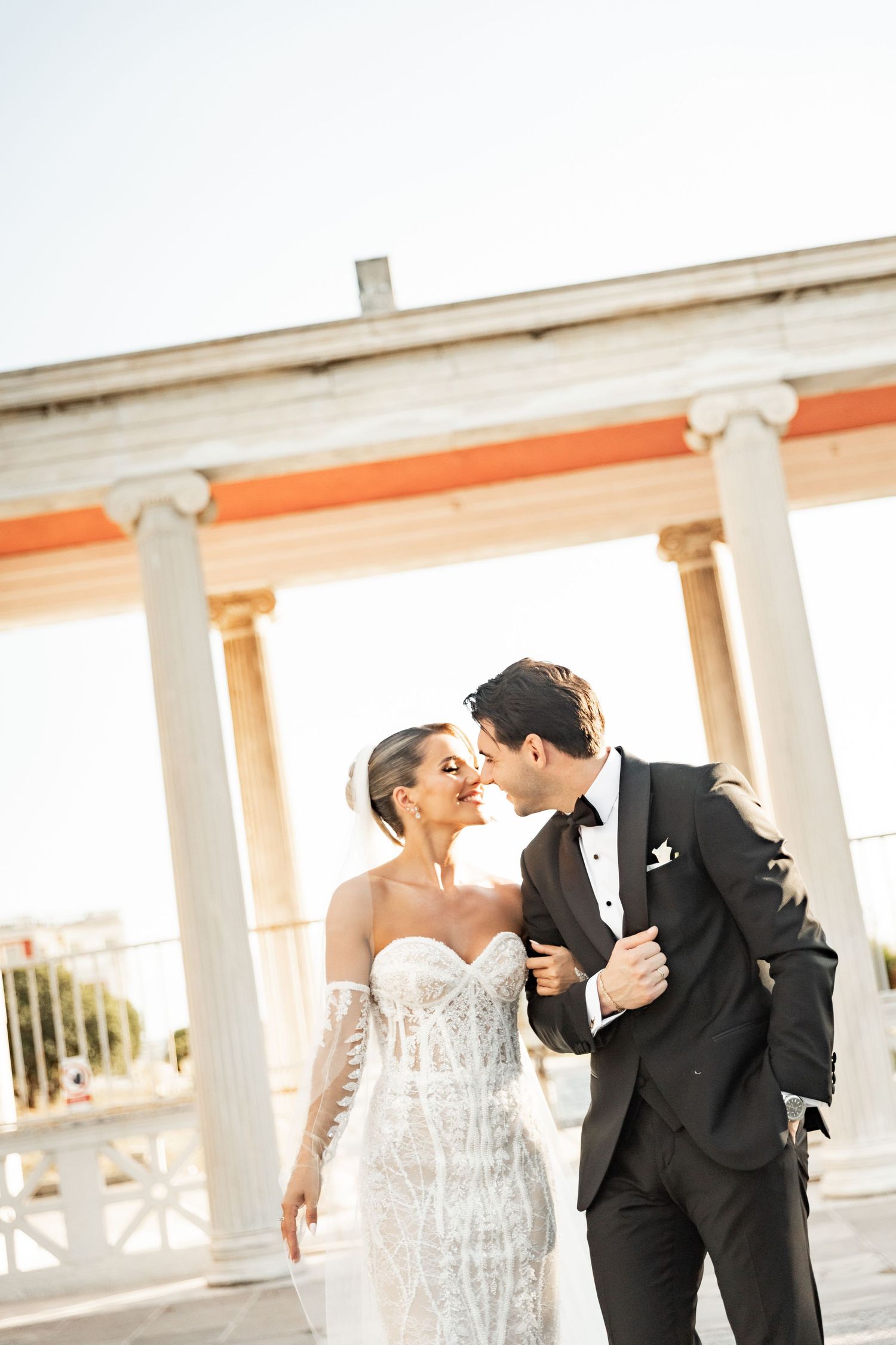 Bride and groom share a romantic moment near white columns in golden sunlight, bride in lace gown, groom in black tuxedo.