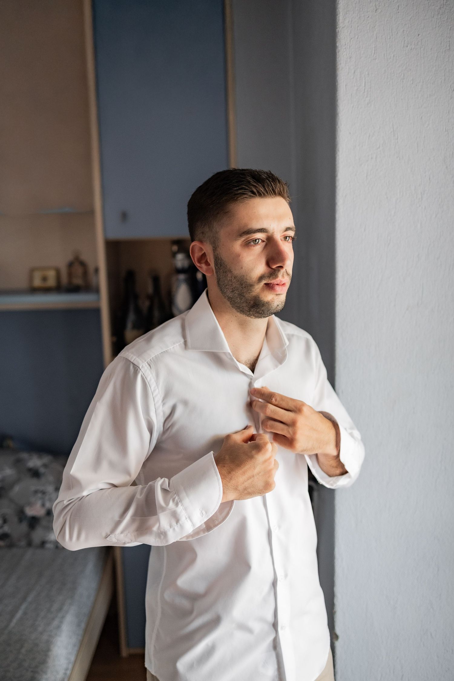 Young man with beard buttoning white dress shirt in bedroom, getting dressed for formal occasion.