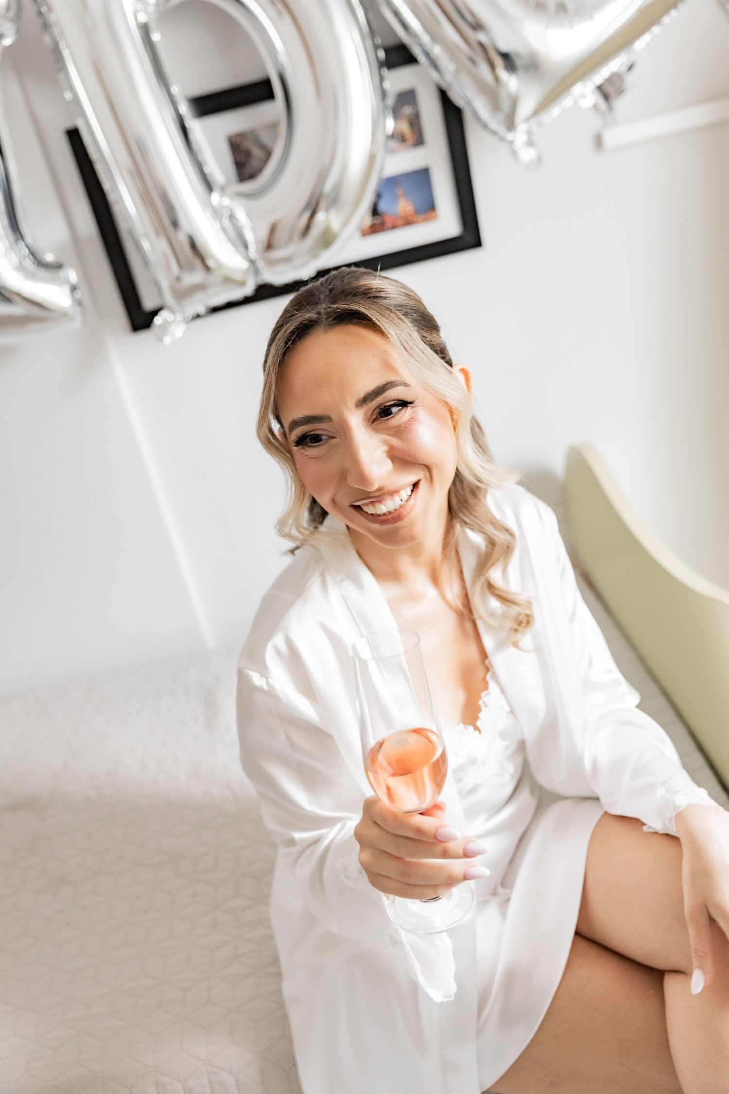 Smiling woman in white robe holding a glass of rosé wine, sitting on a bed with silver balloons in background.