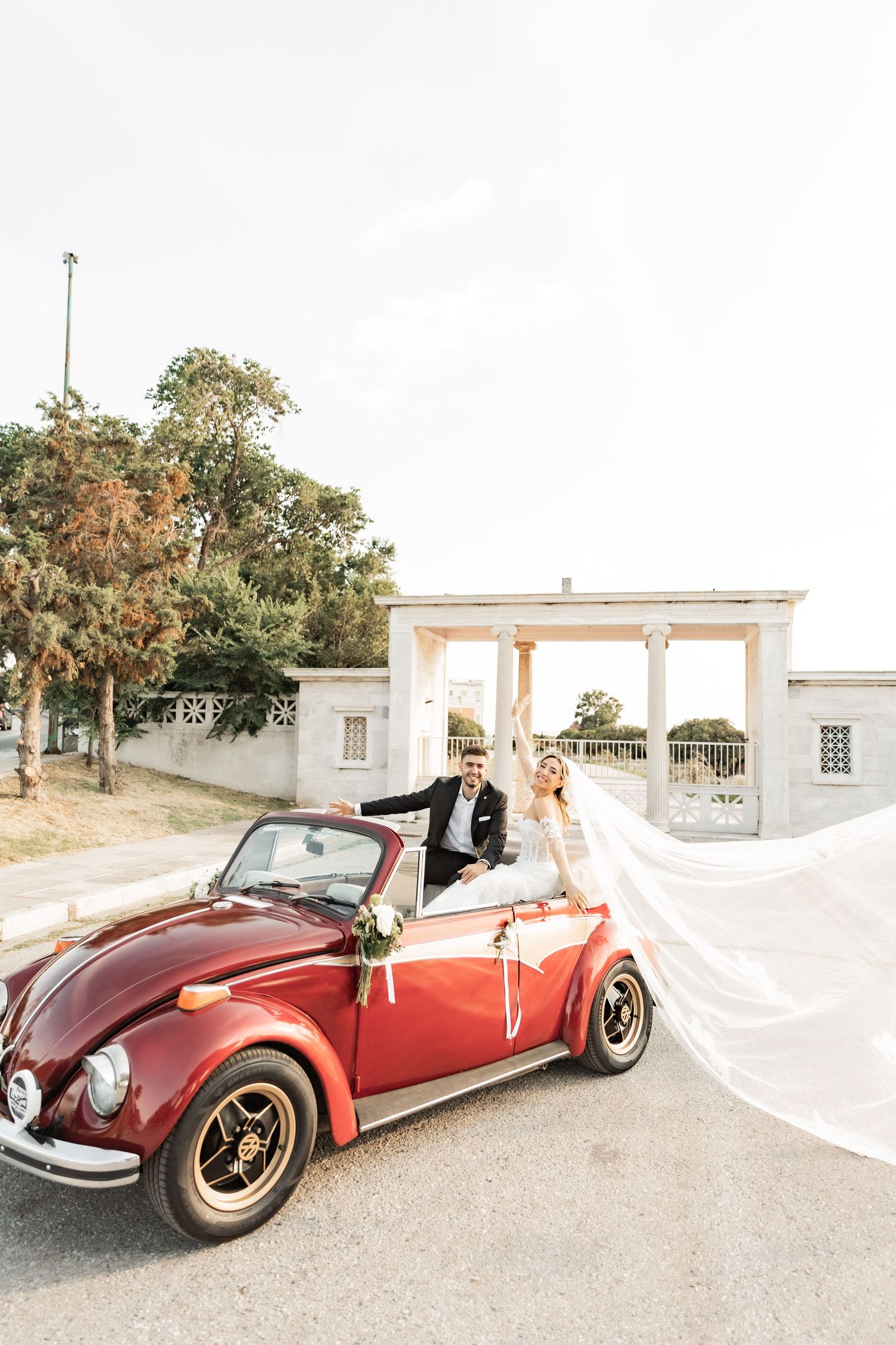 Bride and groom celebrating in a red vintage VW Beetle convertible, with veil flowing in the wind on their wedding day.