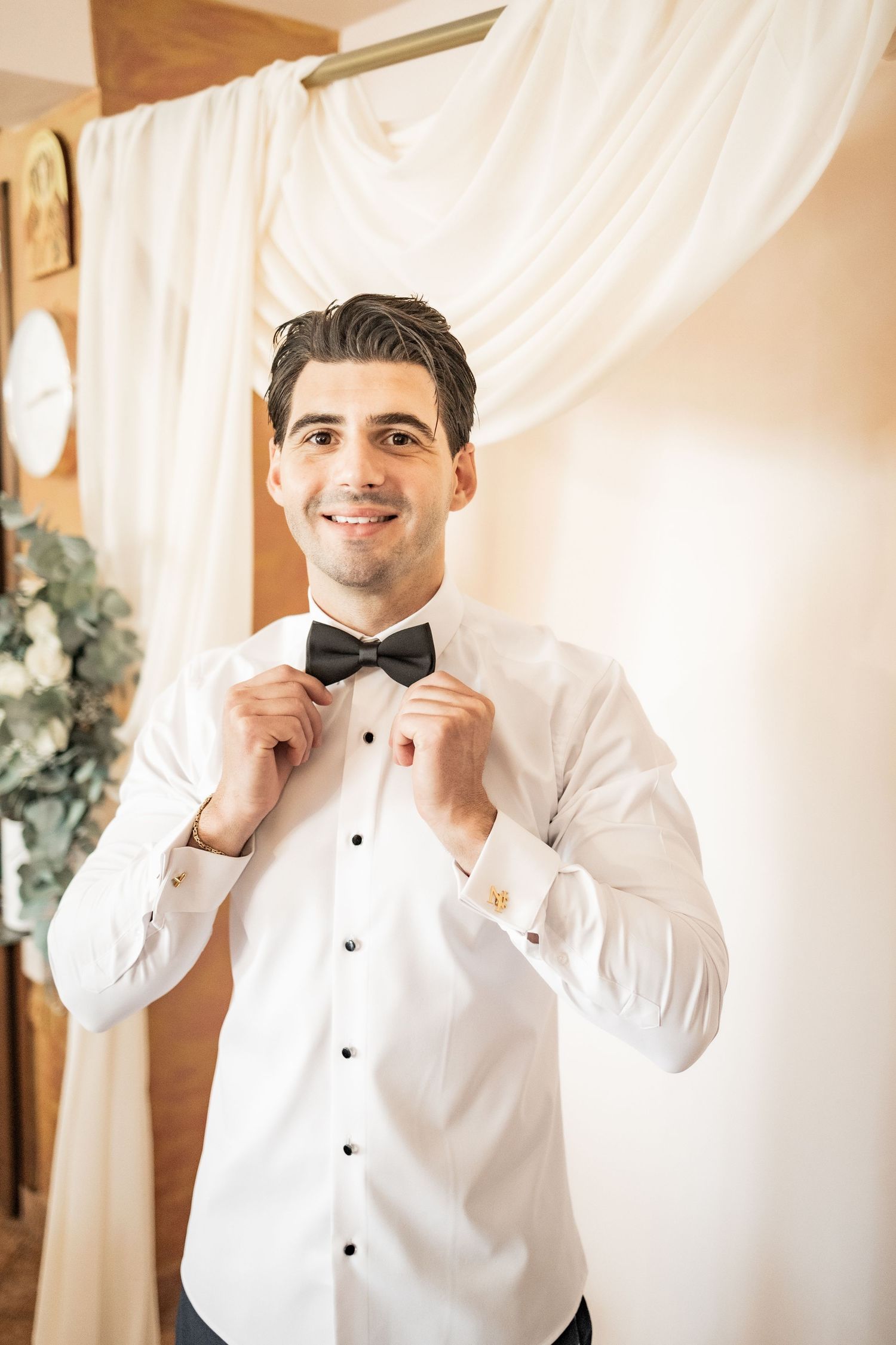 Smiling groom in white tuxedo shirt adjusting black bow tie, getting ready for wedding ceremony.
