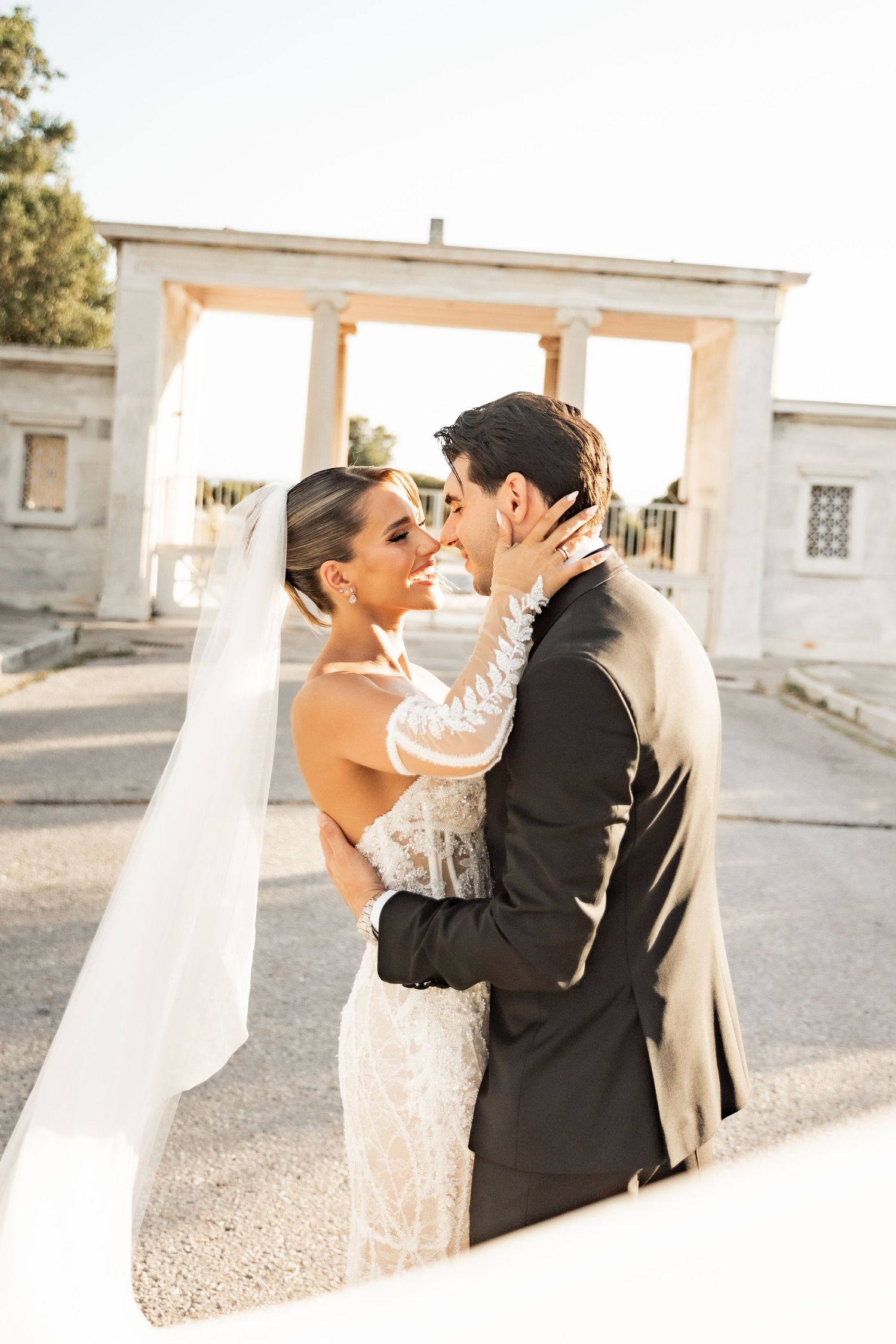 Bride and groom share a romantic moment in front of a classical stone arch, bride wearing lace gown and veil.