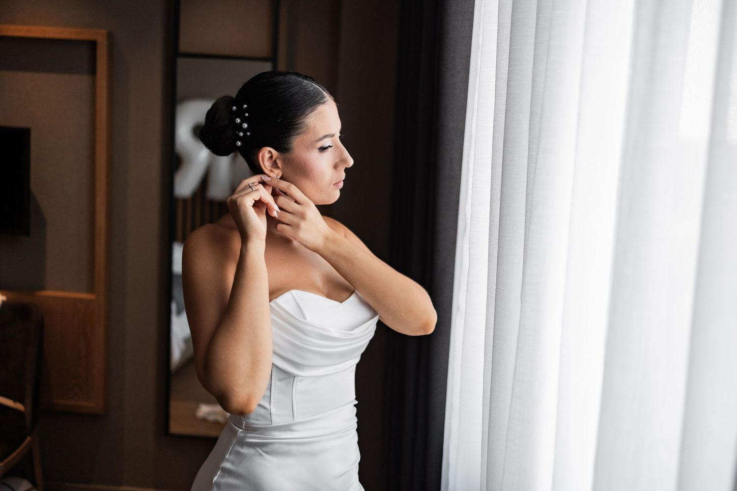 Elegant bride in white dress stands by window in dramatic lighting creating a moody artistic portrait.