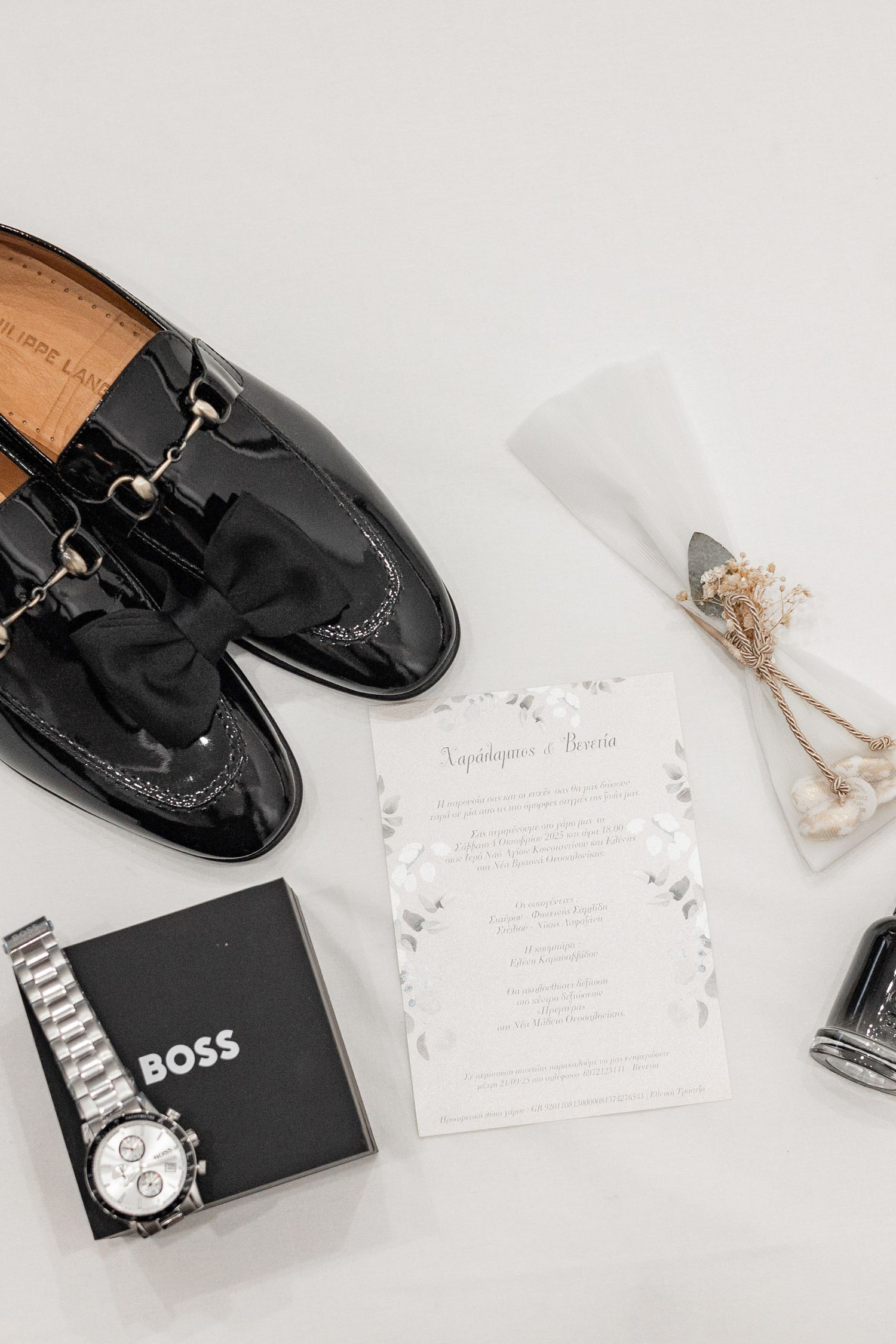 Wedding accessories including black dress shoes, invitations and a Boss wallet arranged on white surface.