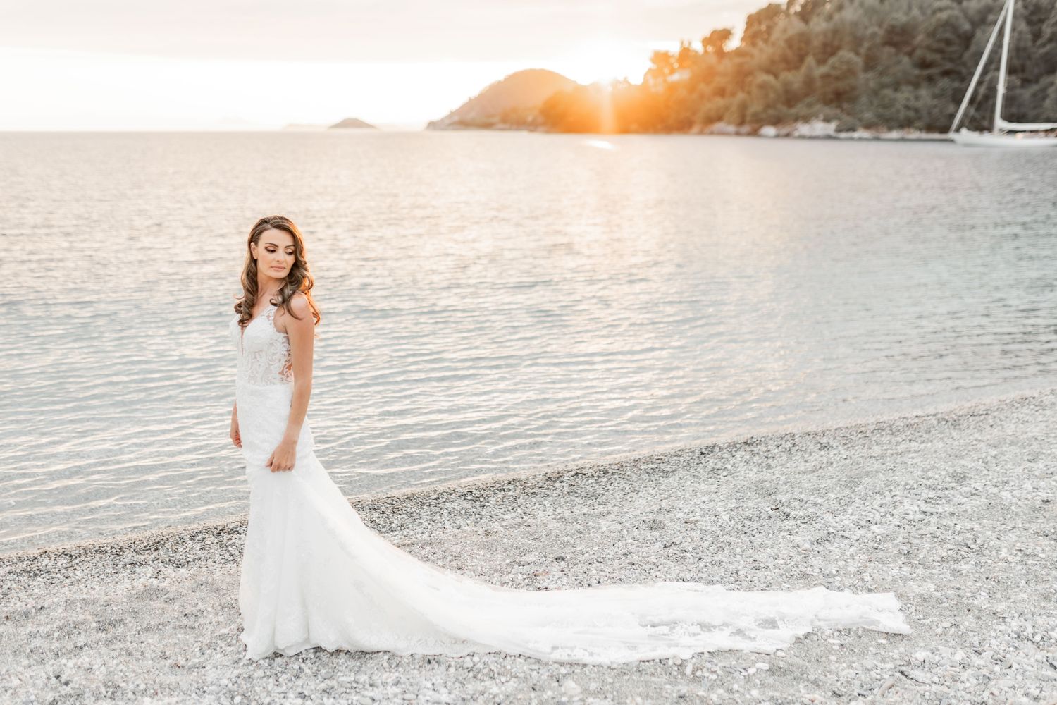Bride in flowing white dress poses on beach at golden hour with ocean view.