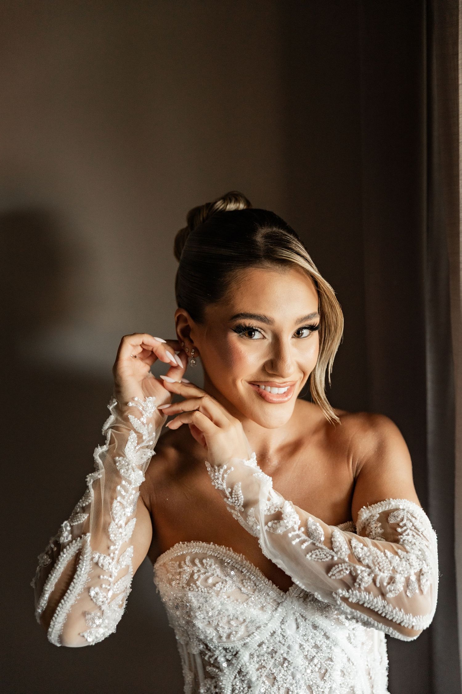 Smiling bride in off-shoulder lace wedding gown with long sleeves putting on earrings, elegant updo hairstyle.