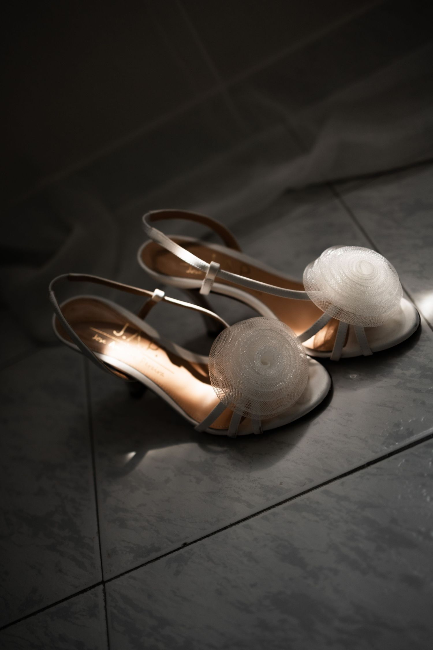 Elegant white bridal sandals with floral organza detail on dark tile floor in soft dramatic lighting.