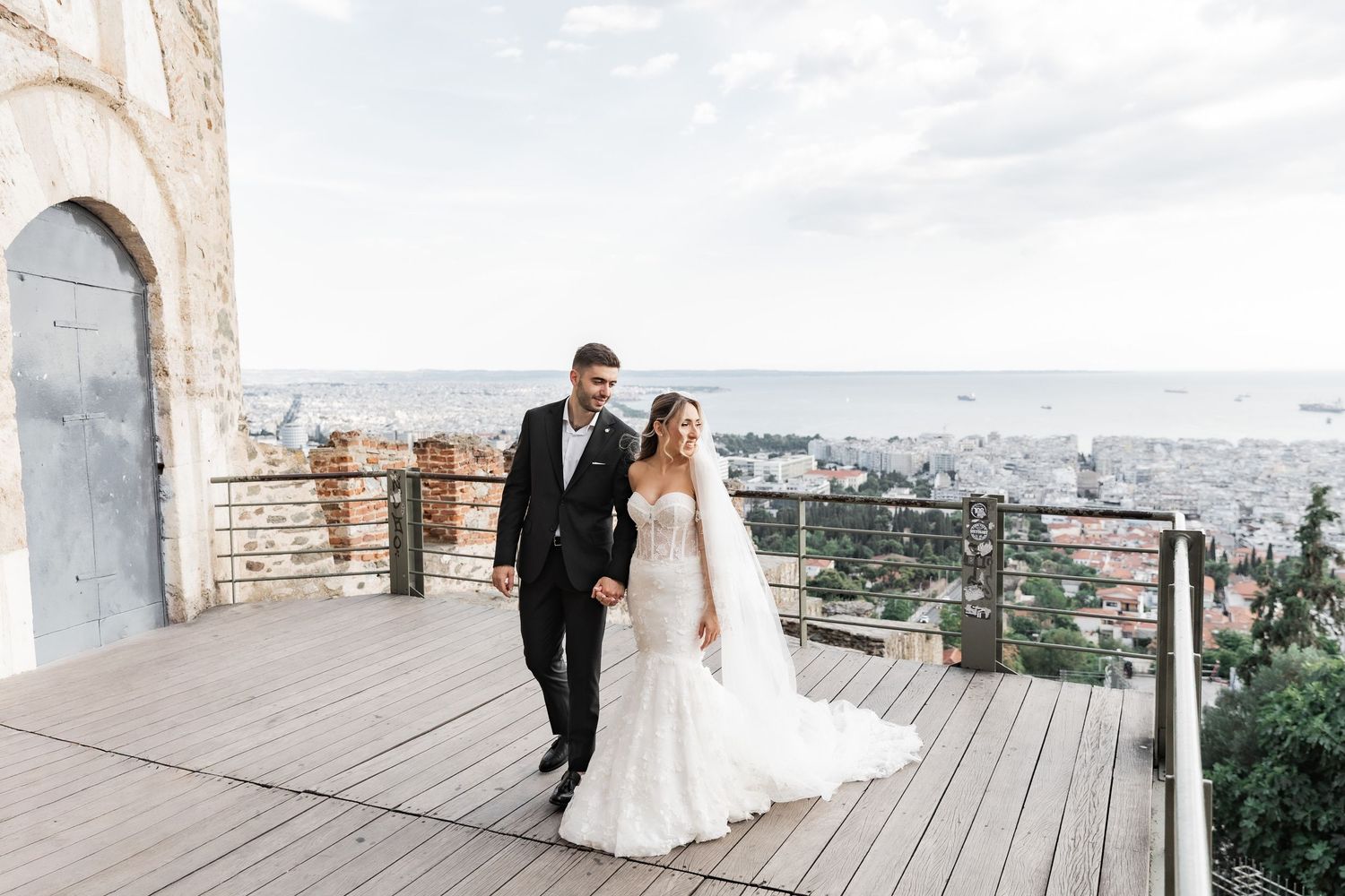 Bride and groom walking hand in hand on a terrace overlooking a coastal city skyline on their wedding day.