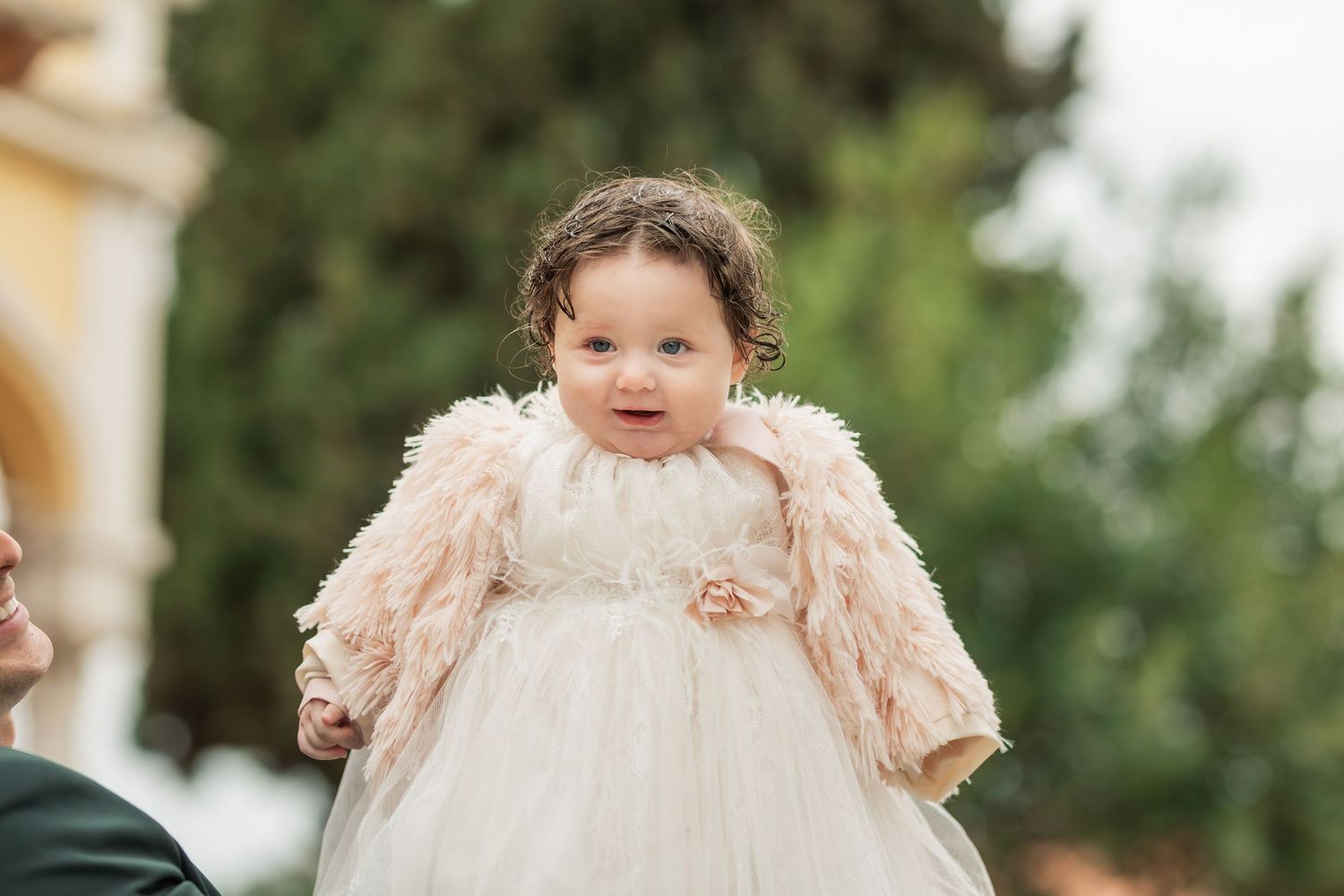 A small child in a cream-colored dress with feathered details stands in a garden setting.