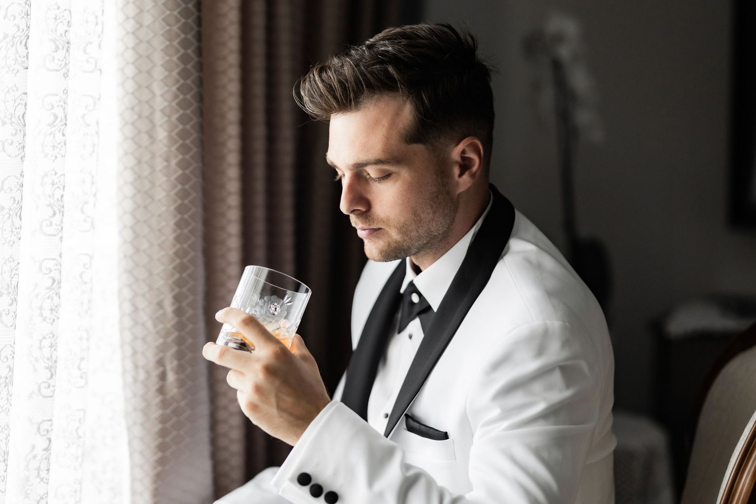 A groom in a white tuxedo jacket and black bow tie sips from a glass in a dimly lit room.
