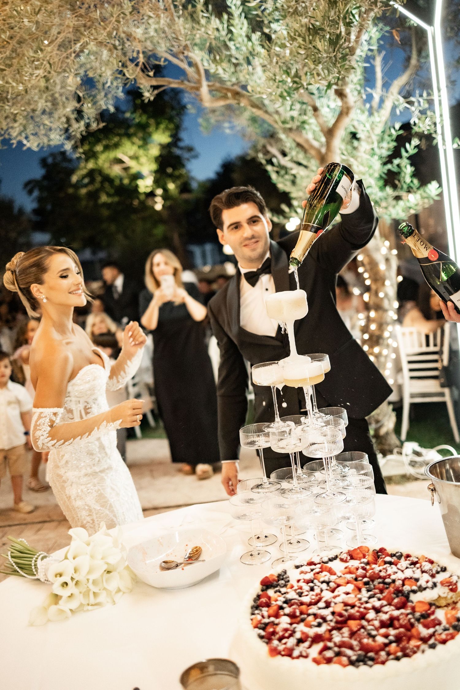 Bride and groom pouring champagne tower at elegant outdoor wedding reception at night with guests celebrating.