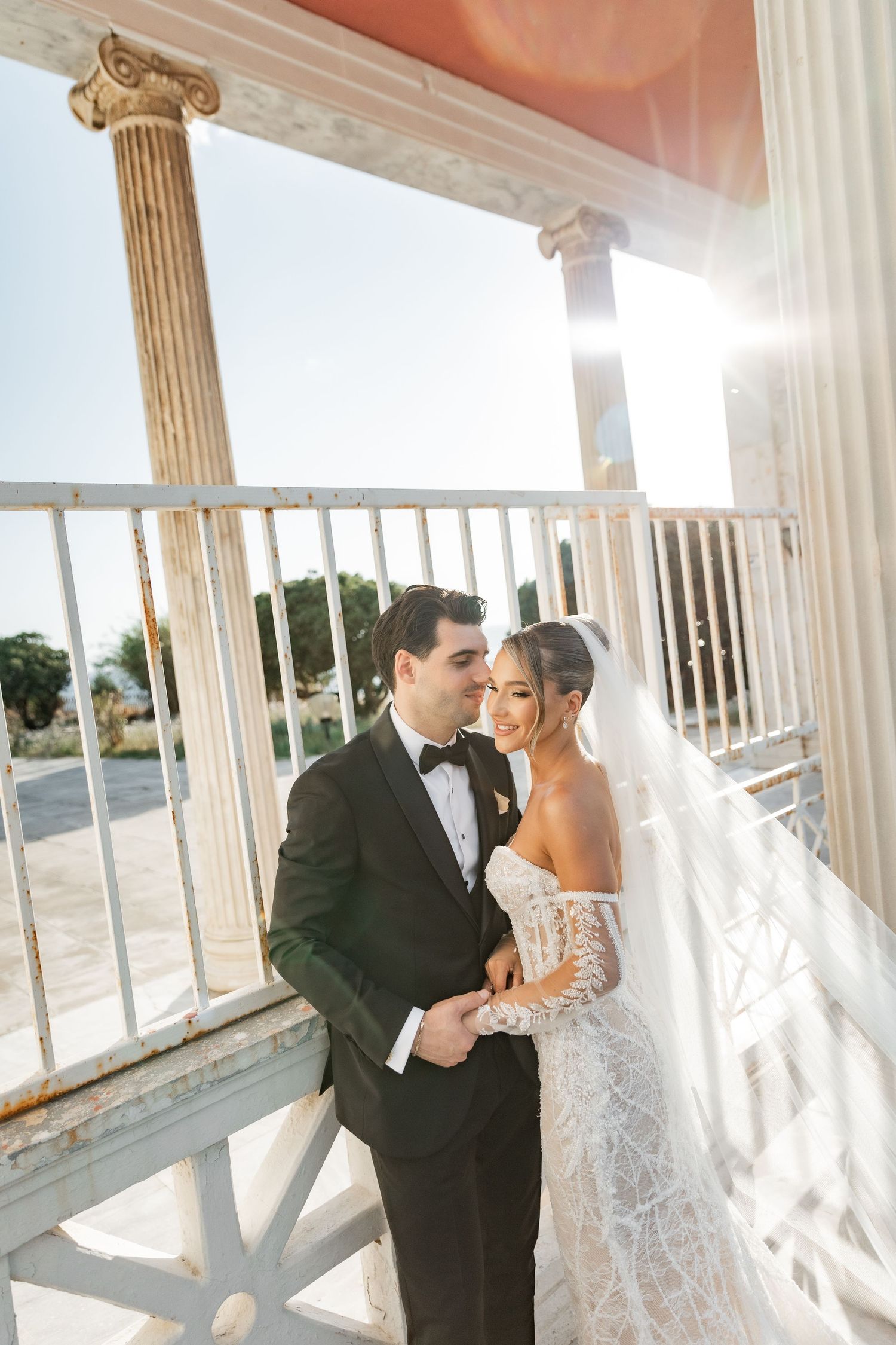 Bride and groom share a romantic moment on a sunlit classical colonnade, she in lace gown with veil, he in black tuxedo.