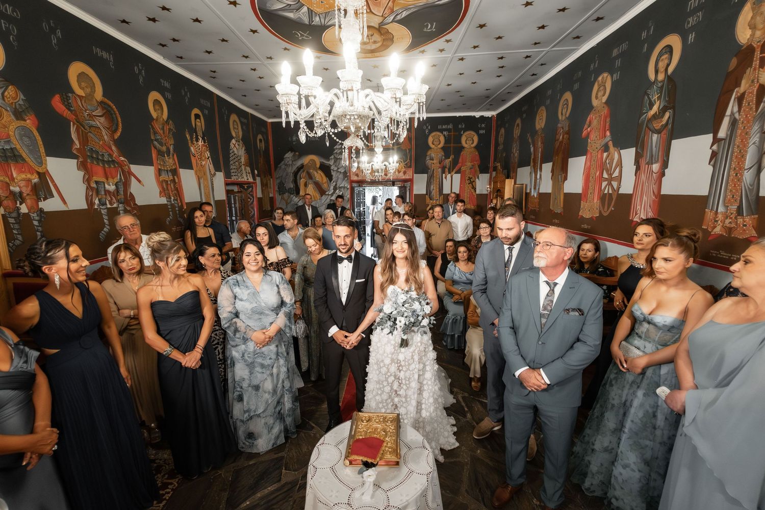 Wedding guests gather in an elegant ballroom with ornate chandeliers and decorative wall murals.