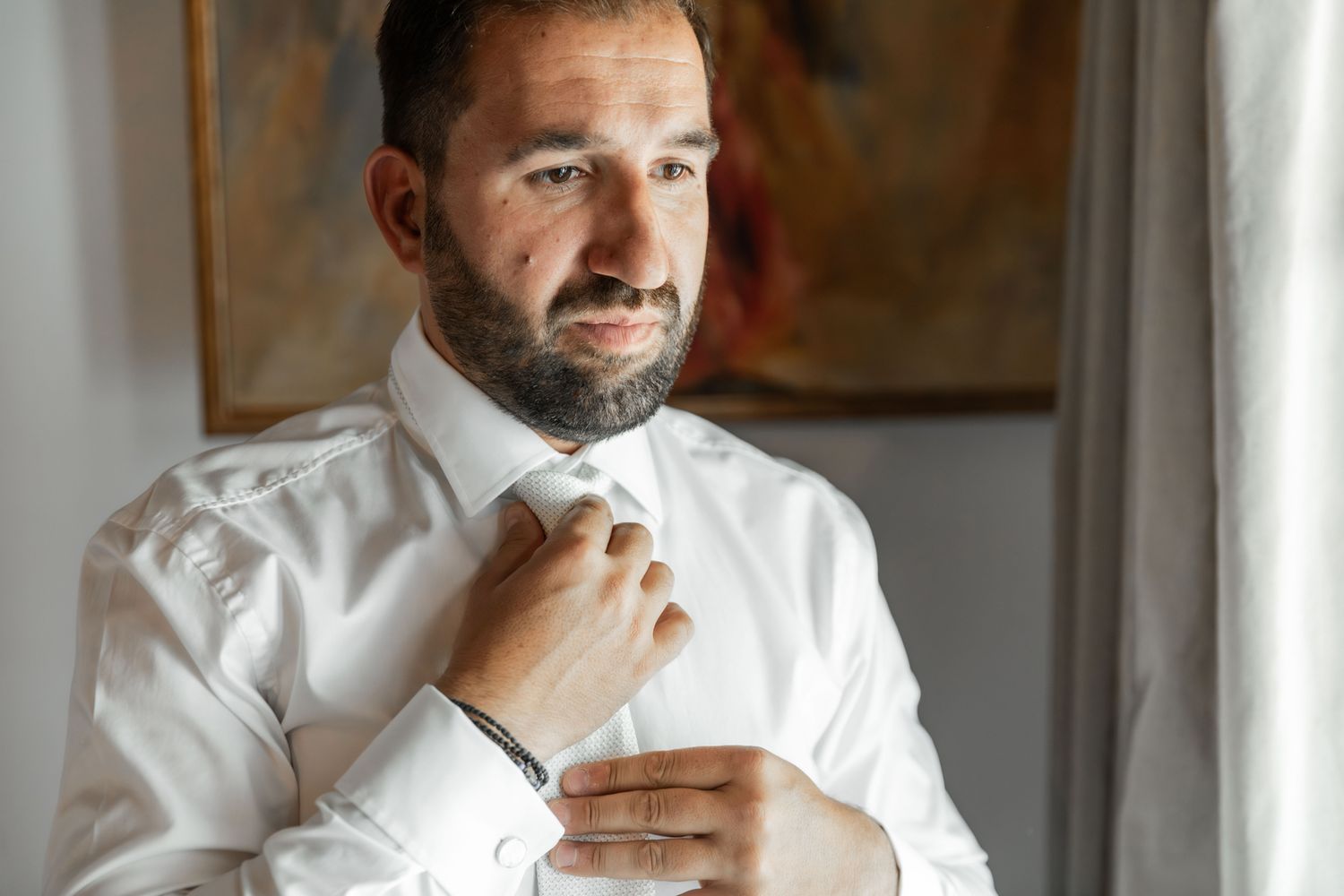 A person wearing a white shirt stands thoughtfully by a window in natural light.