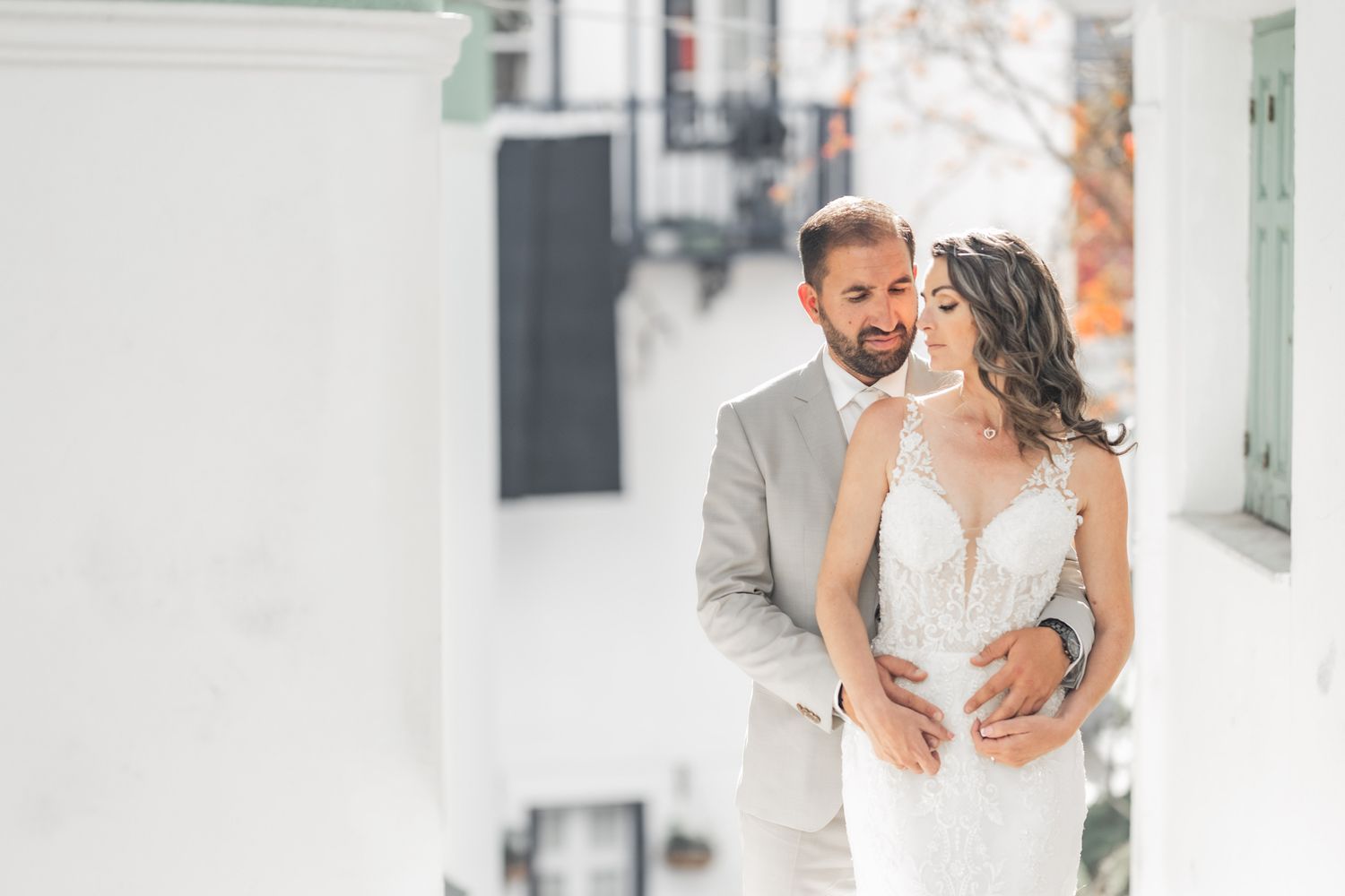 Romantic wedding couple embraces in soft natural light against white walls.