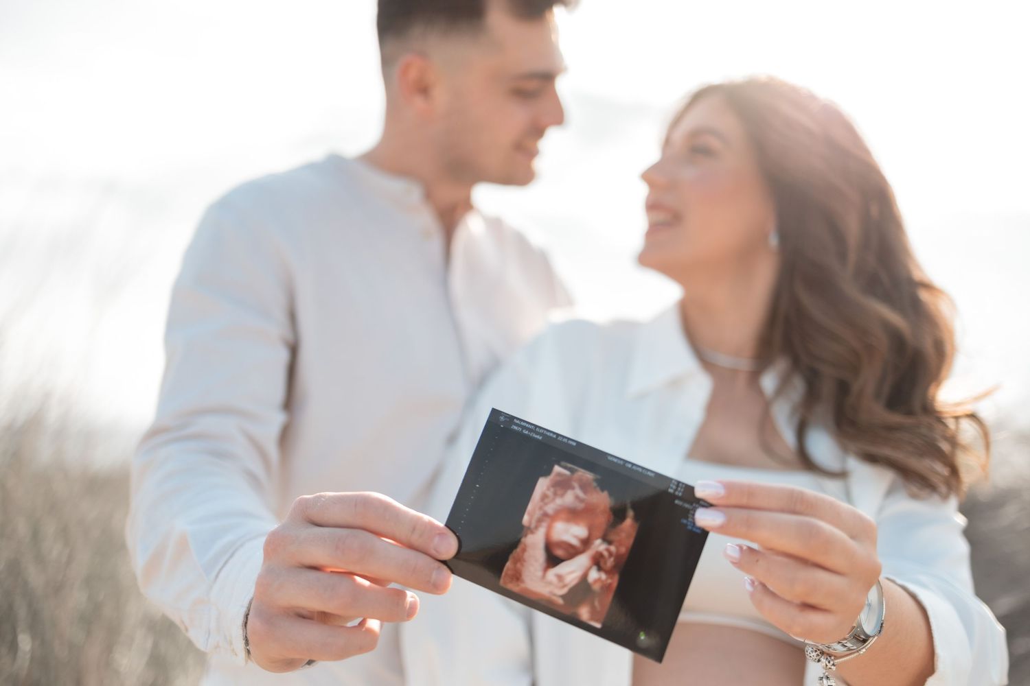 A couple outdoors holds up a polaroid photo while smiling at each other in bright sunlight.