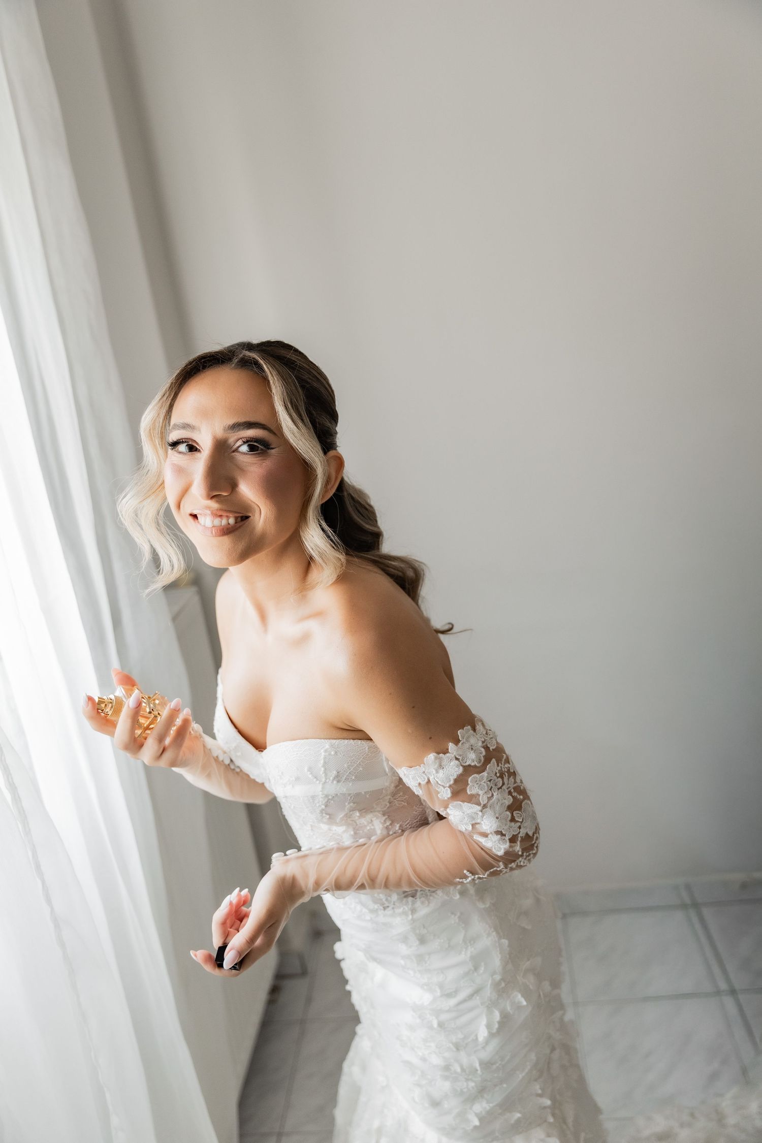 Smiling bride in strapless lace wedding gown with floral sleeves poses by a bright window in soft natural light.
