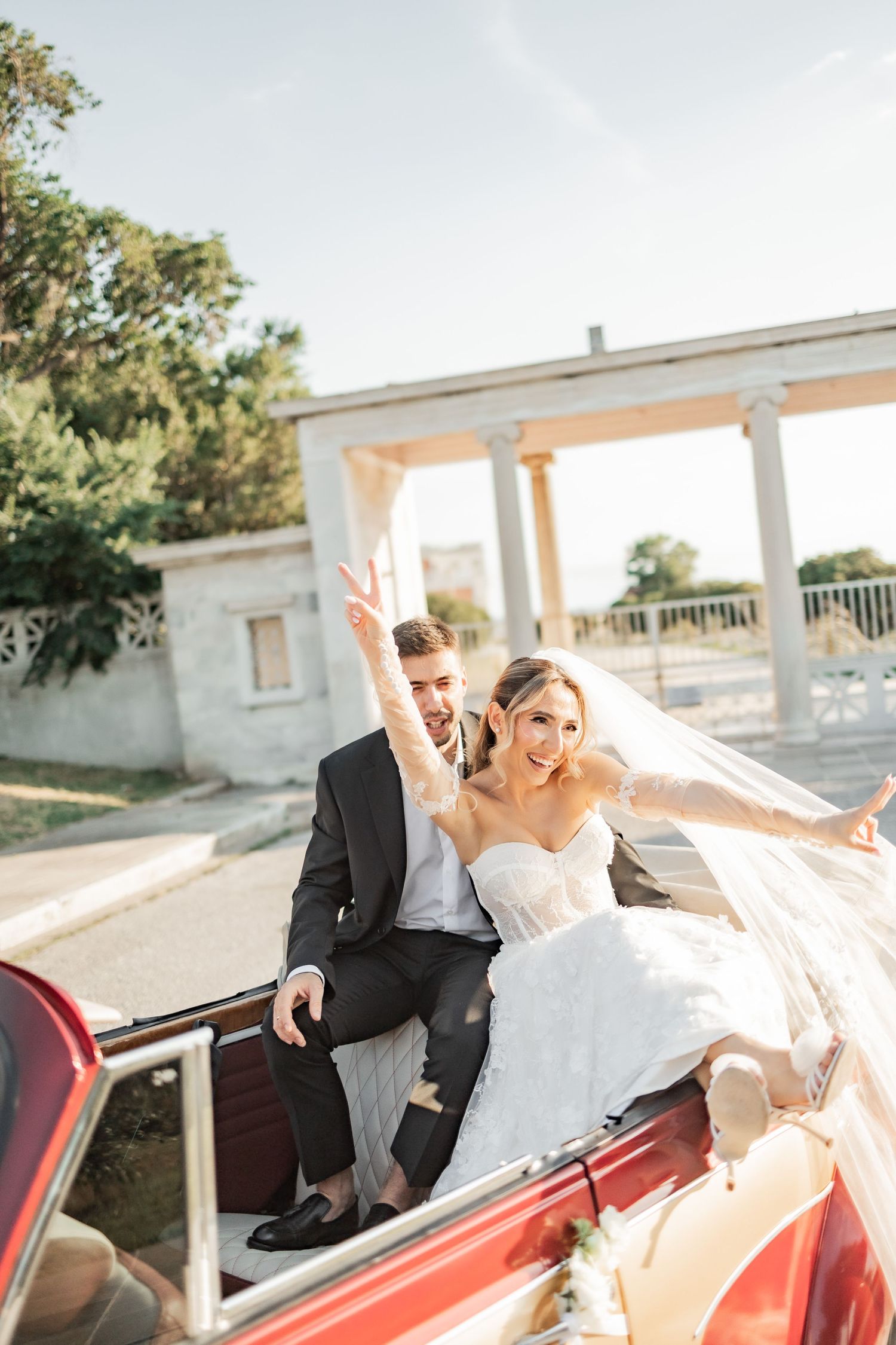 Joyful newlyweds celebrating in a vintage red convertible car, bride in white gown with veil, groom in dark suit, making peace sign.