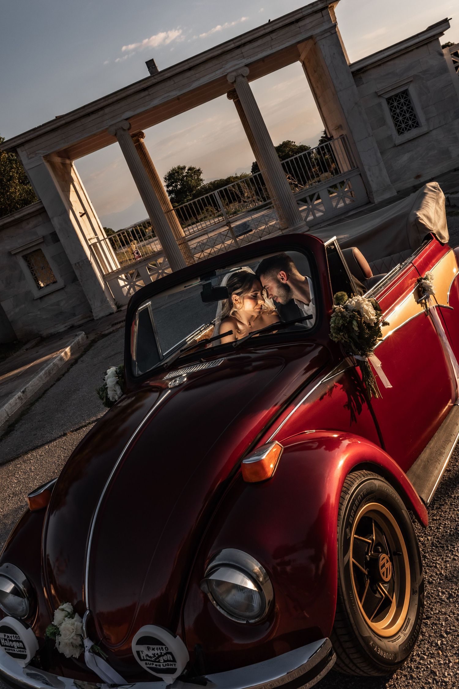 A couple shares a romantic kiss in a vintage red VW Beetle convertible adorned with flowers outside an elegant venue.