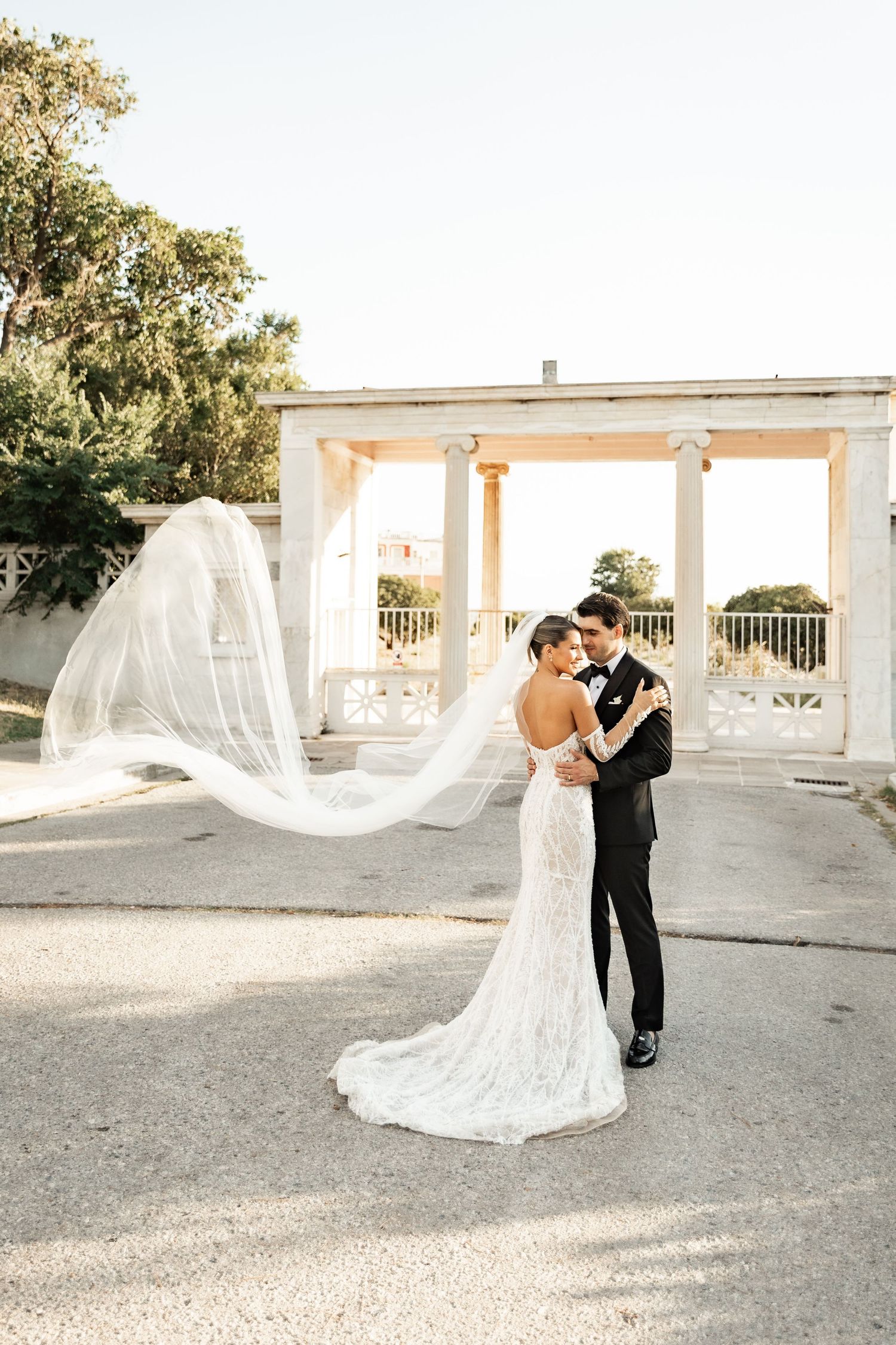 Bride in lace gown with flowing veil embraces groom in black tuxedo before elegant white columned pavilion.