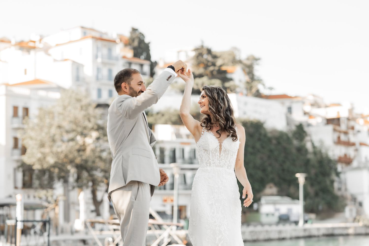 Wedding couple dances by Mediterranean waterfront with white buildings in background.