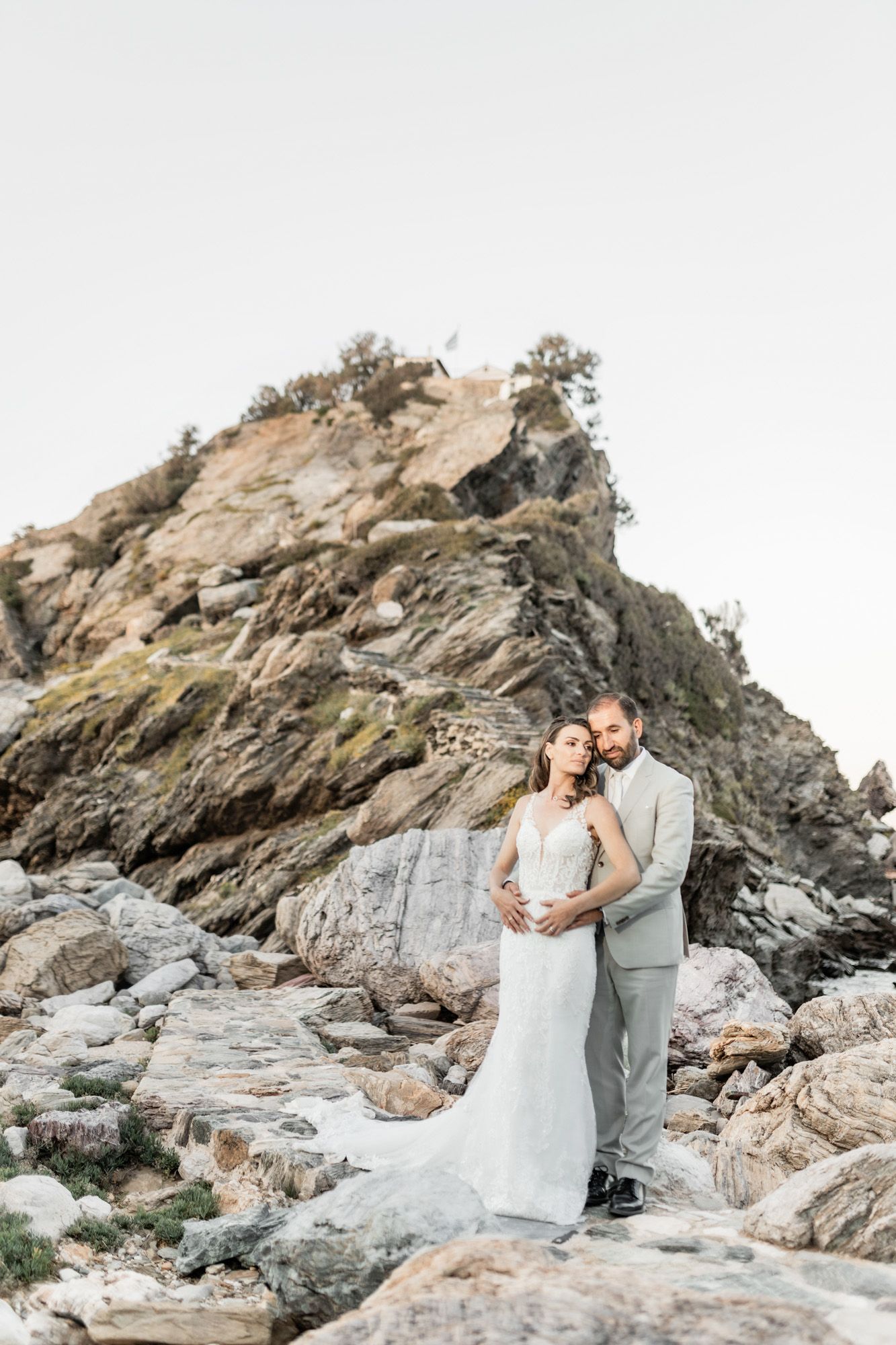 A wedding couple poses against dramatic rocky cliffs on a coastal landscape at sunset.