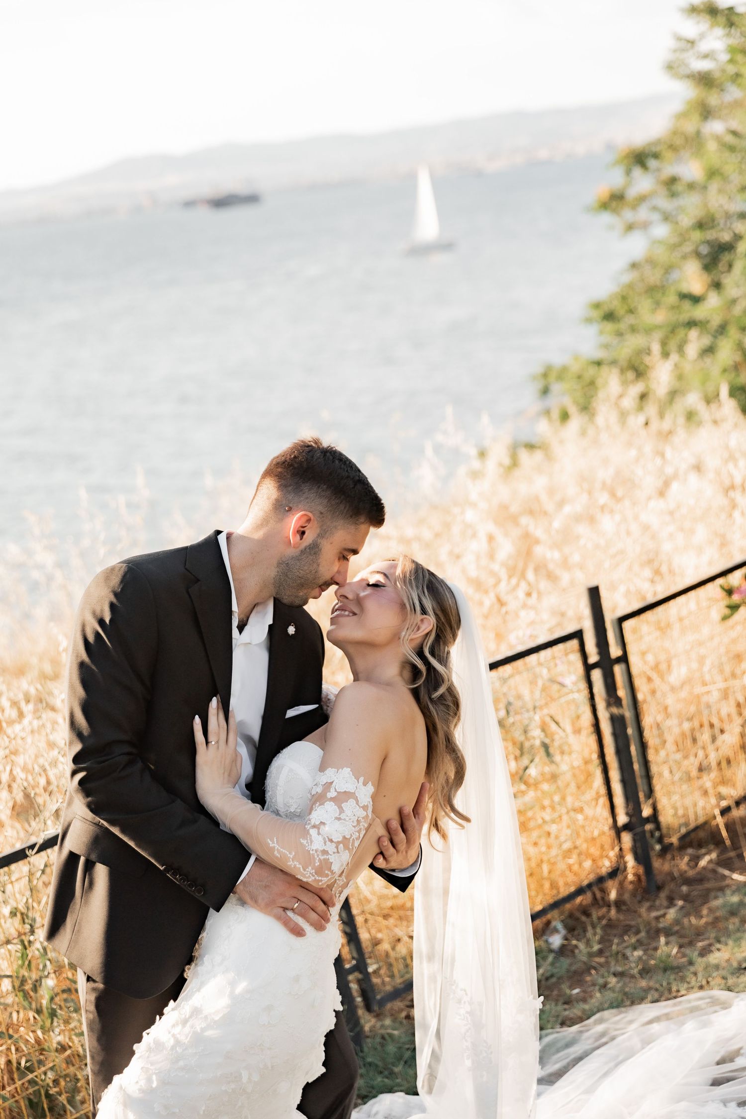 A bride and groom share a romantic moment outdoors near golden hills and a bay with a sailboat in the background.