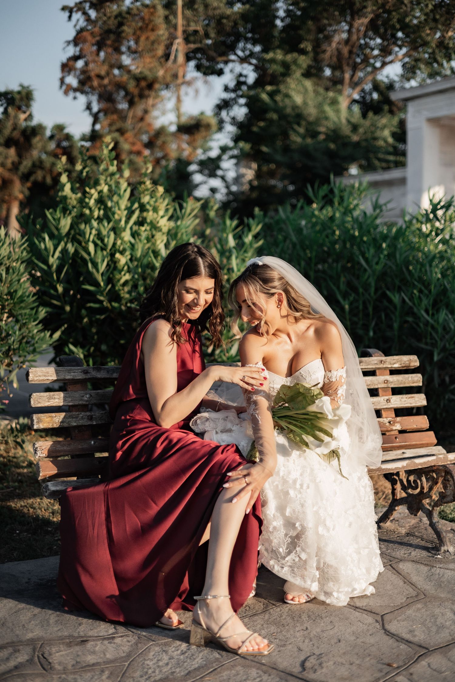 Bride in white gown and bridesmaid in red dress sitting on bench, smiling and looking at wedding ring outdoors.