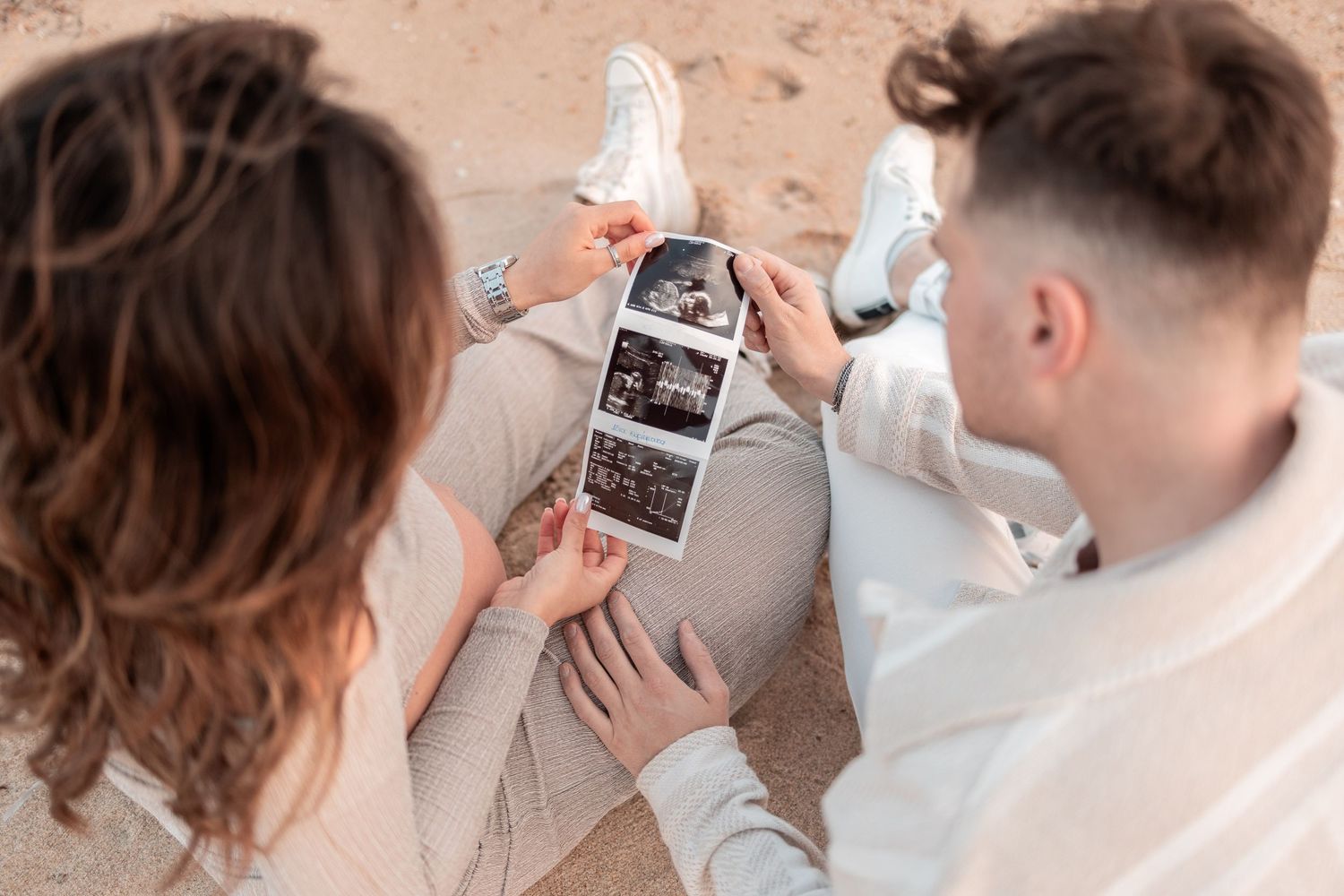 Couple sitting together looking at ultrasound photos on a phone while wearing casual white clothing.