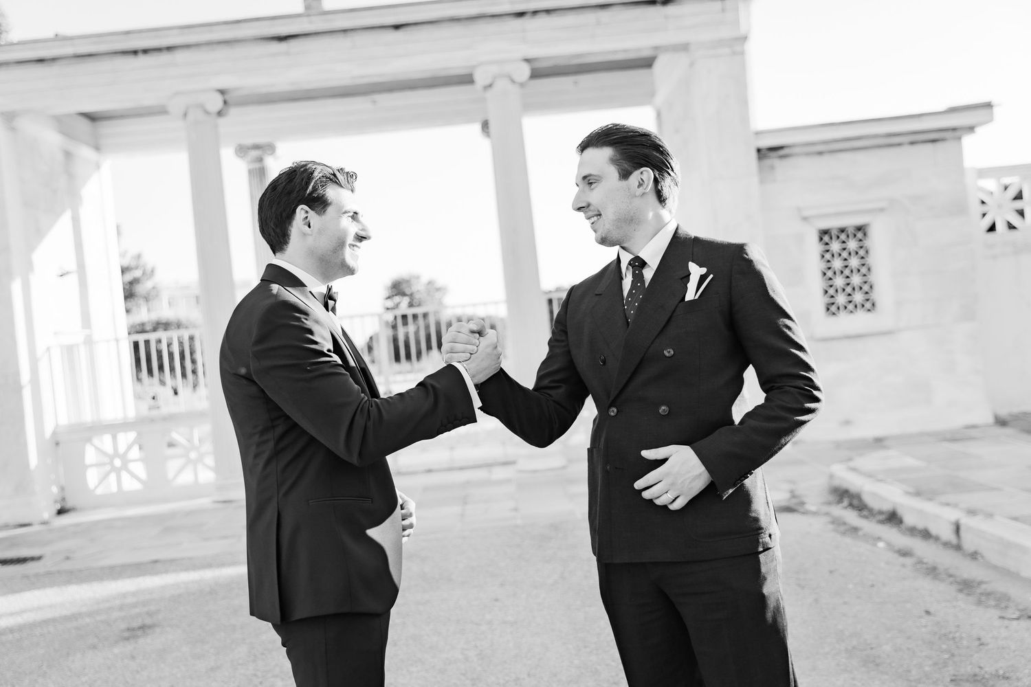 Two grooms in black suits share a joyful moment shaking hands at an elegant outdoor wedding venue.