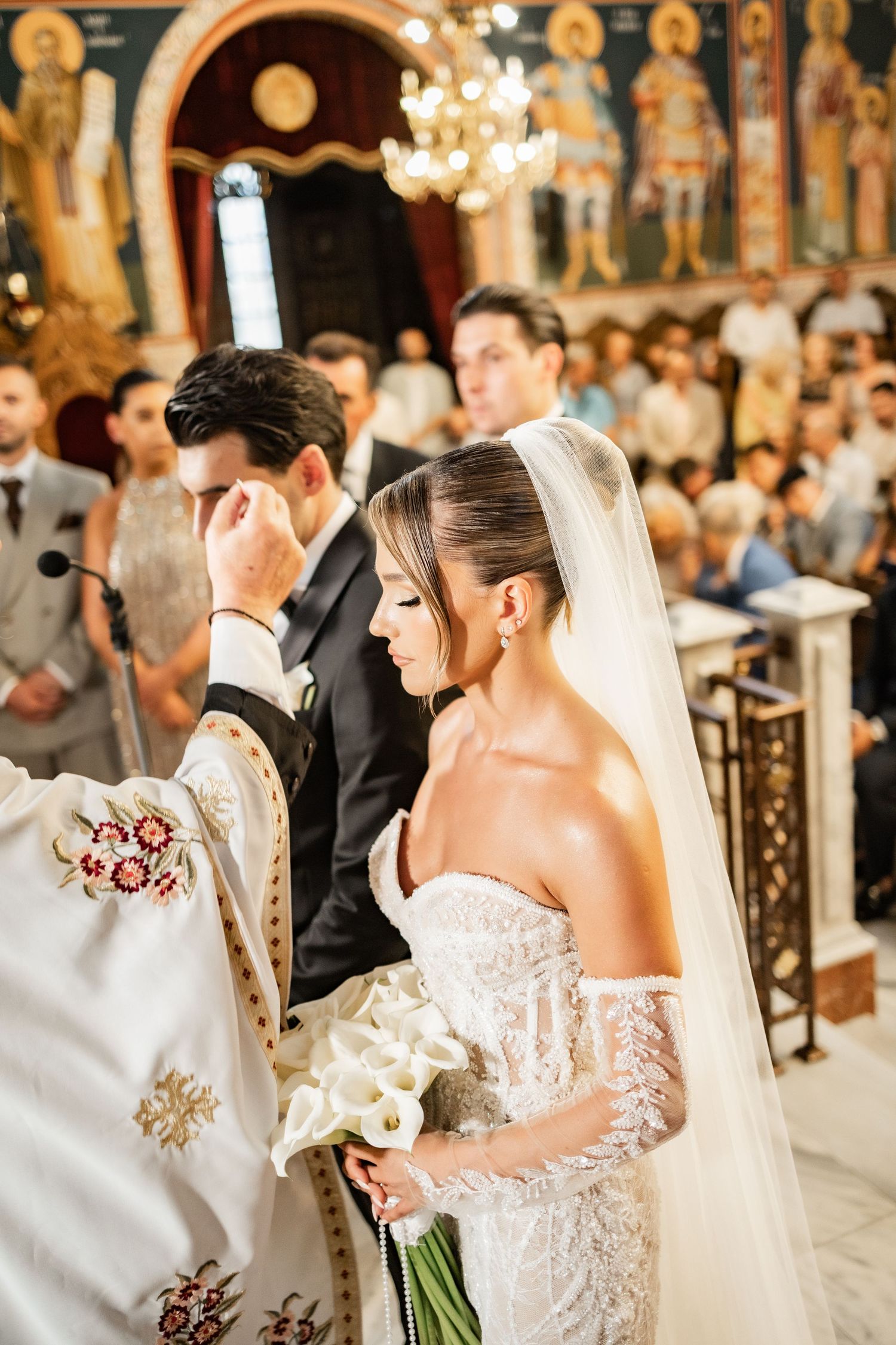 Bride and groom stand before an Orthodox priest during a church wedding ceremony, bride holding white calla lily bouquet.