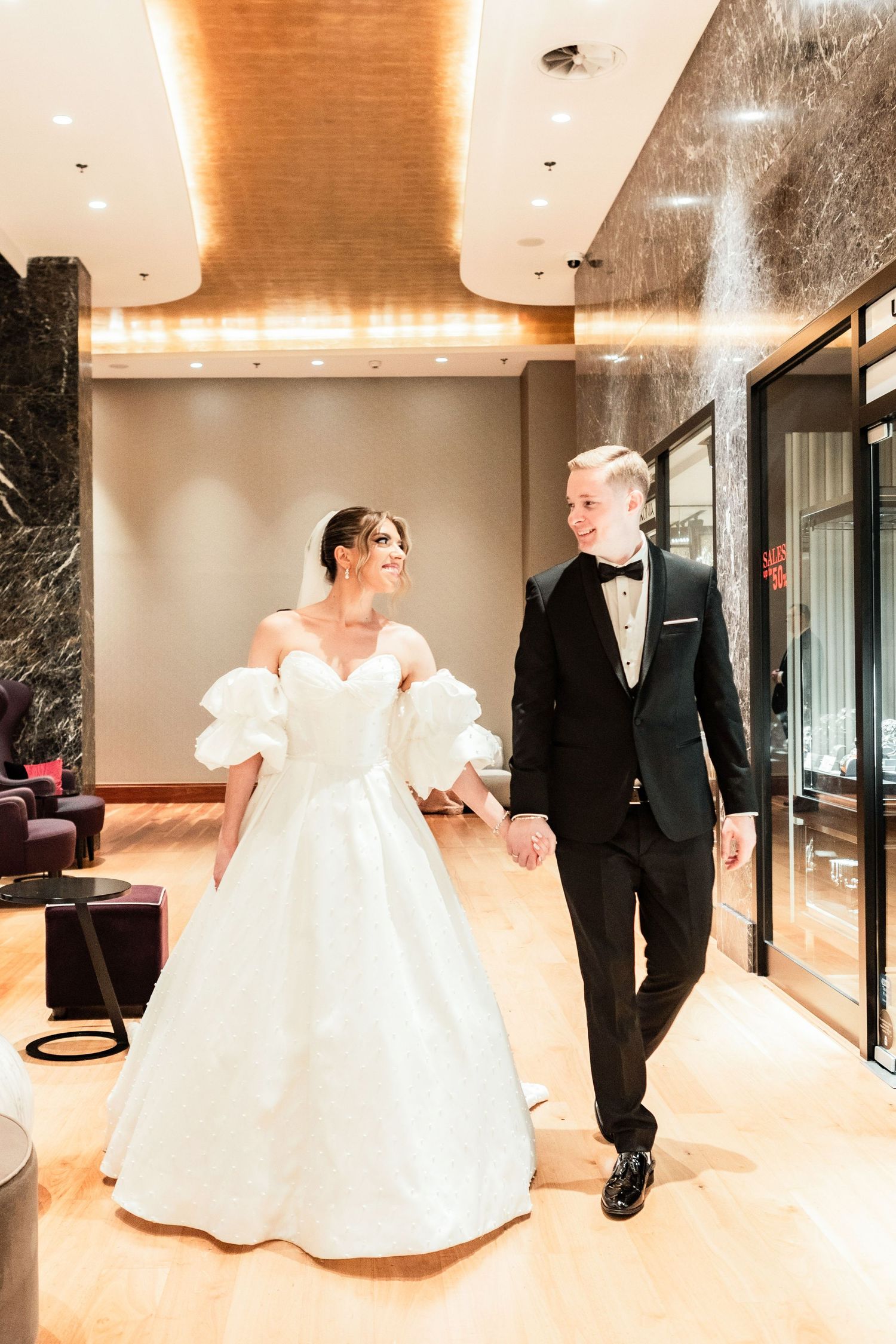 Wedding couple walking through a modern hallway with marble walls and warm lighting.