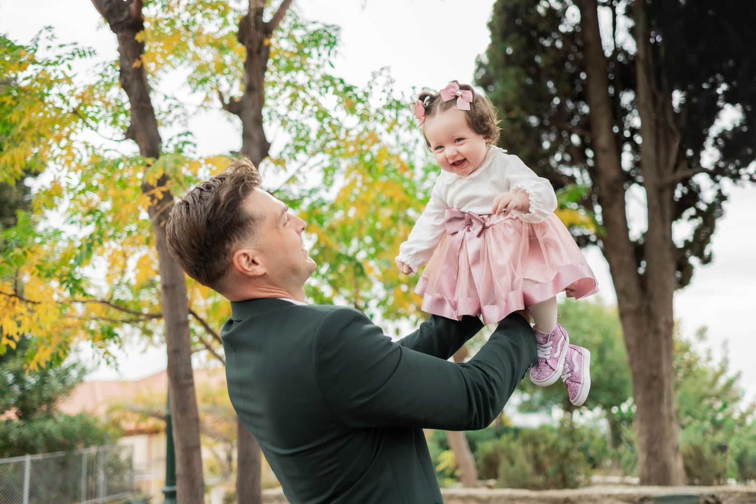 A parent plays with a small child wearing pink in an outdoor autumn setting with trees in the background.