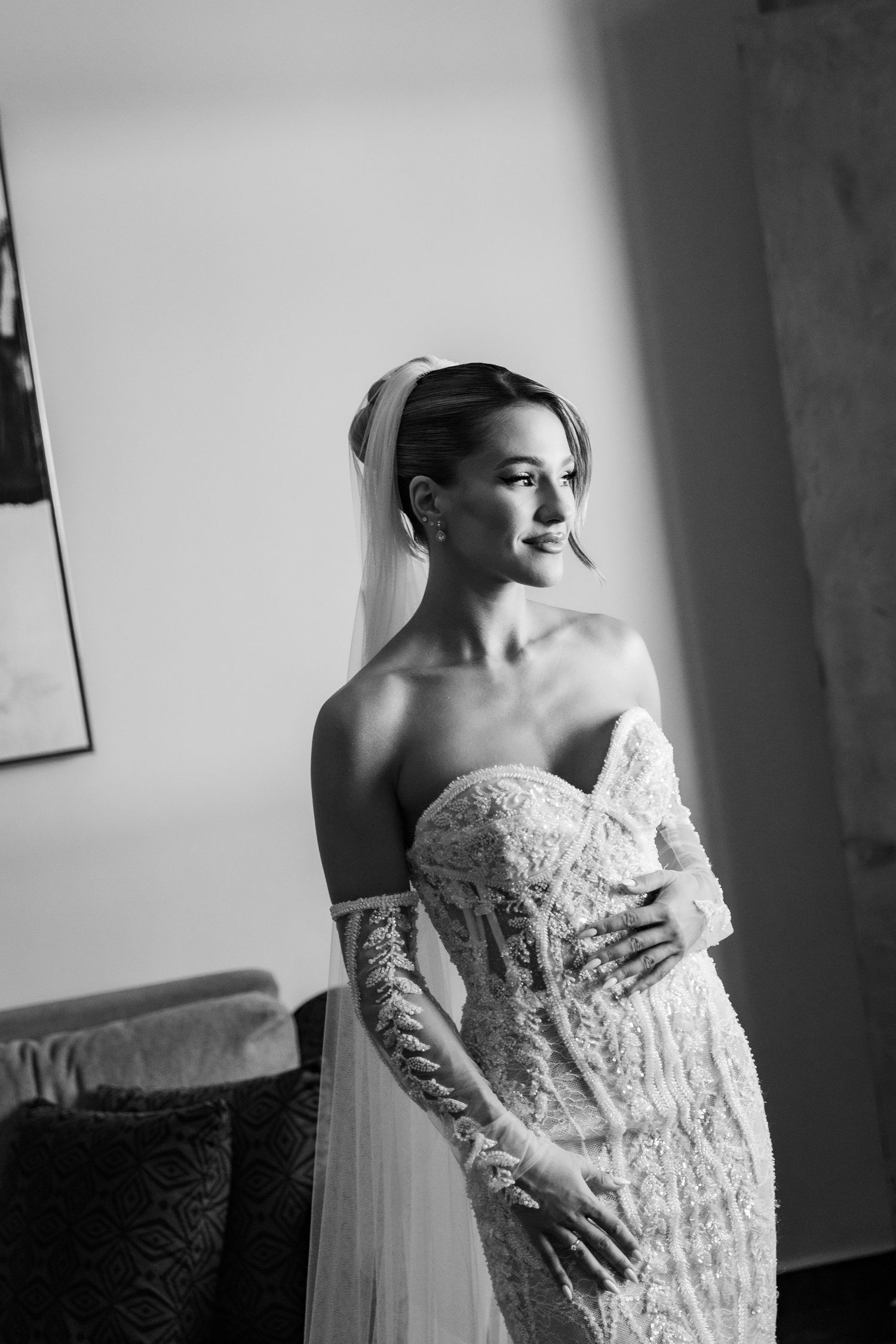 Black and white portrait of an elegant bride in a lace strapless gown with veil, posing gracefully indoors.