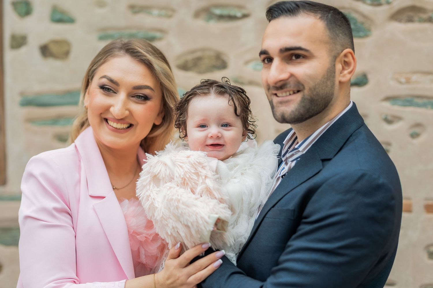 A family poses together against a stone wall, with parents wearing pink and navy blazers and a baby in white lace.
