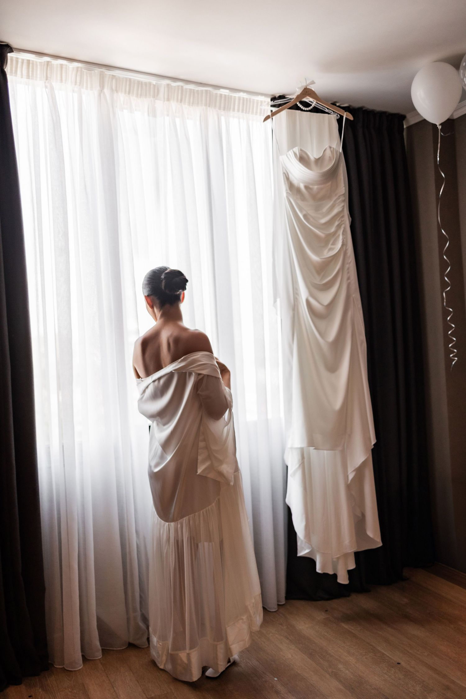 Artistic silhouette of wedding dress preparation with gown hanging by window.