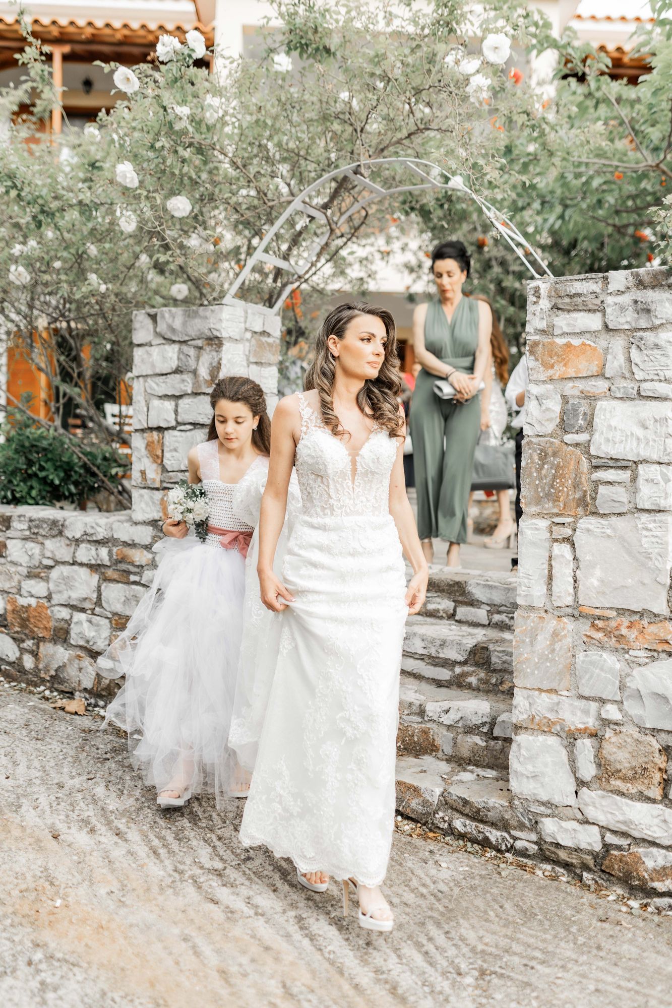 Bridesmaids in sage green dresses walk along a stone wall pathway during wedding festivities.