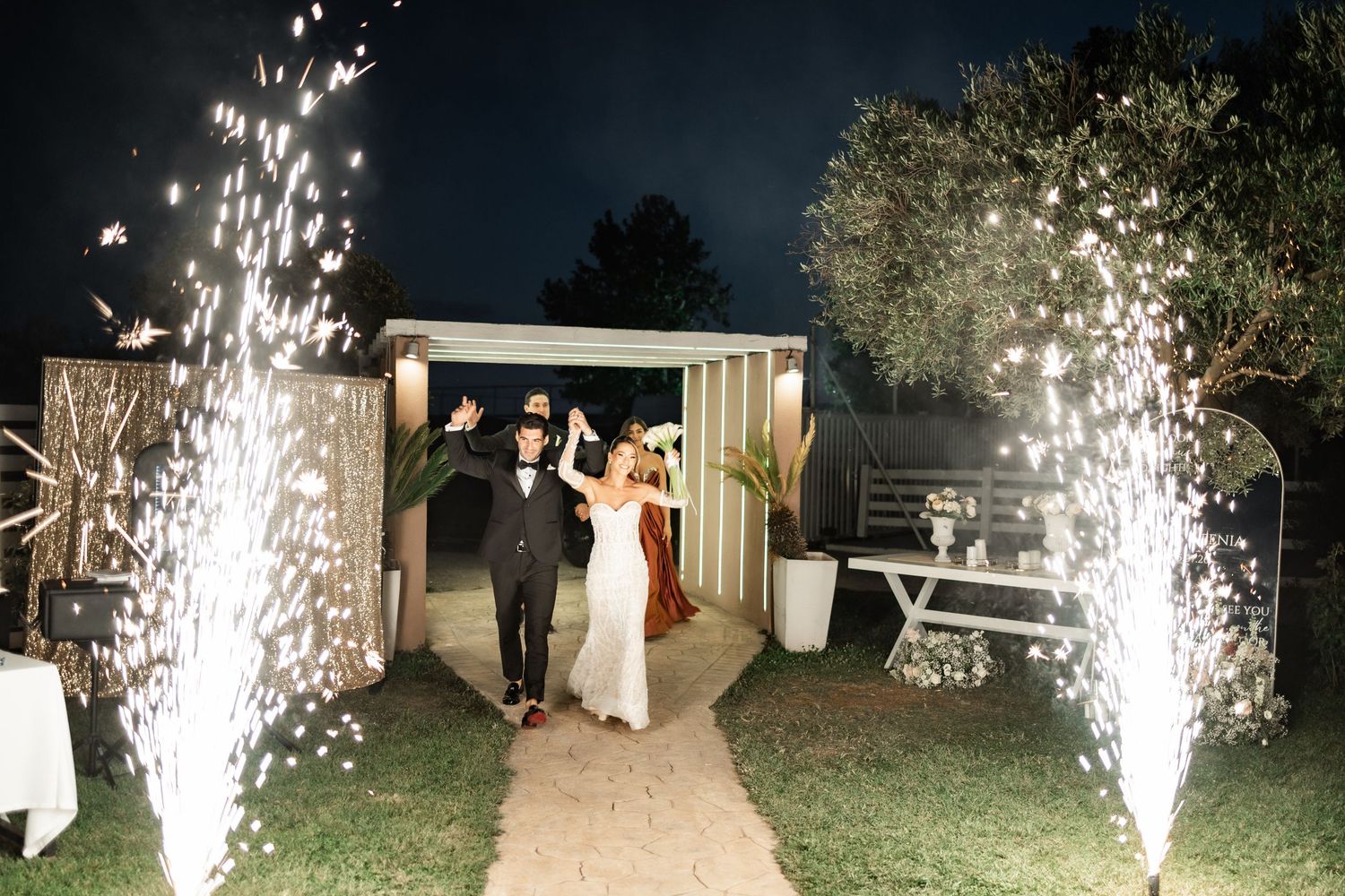 Bride and groom walk through sparkling cold fireworks display at night during outdoor wedding reception celebration.