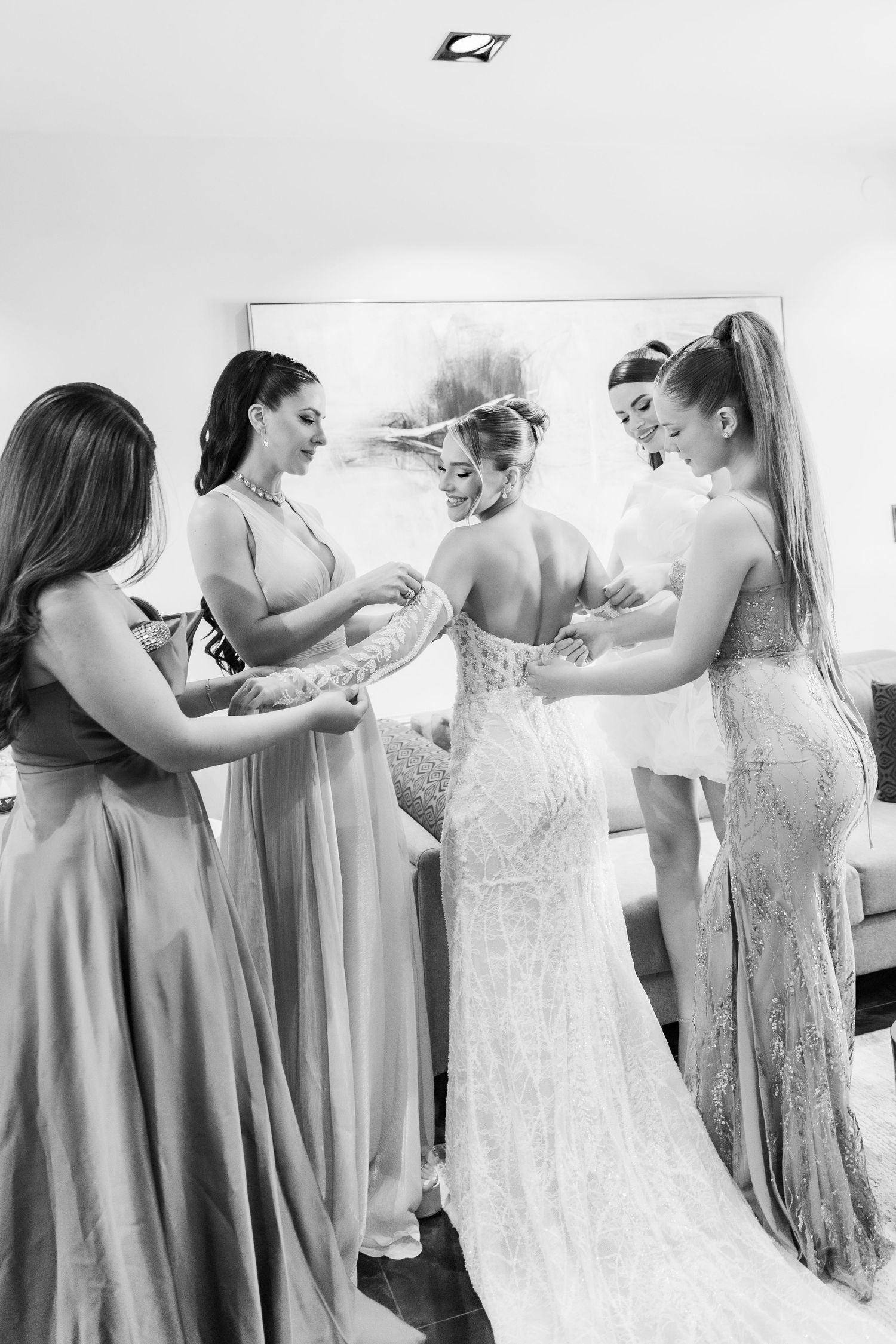 Bridesmaids in elegant gowns help bride with lace wedding dress in black and white getting ready photo.