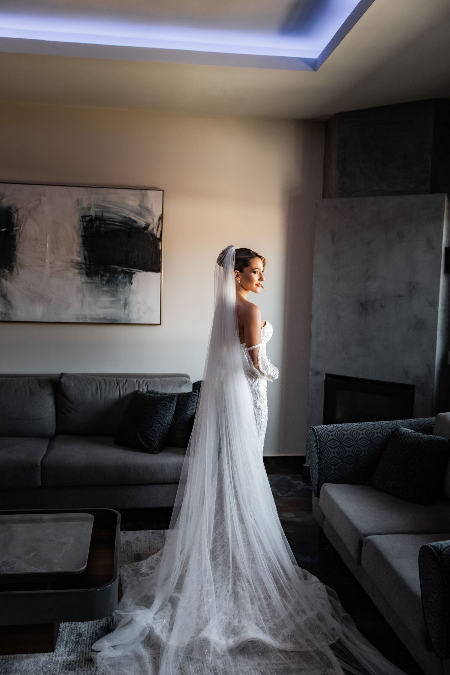 Bride in elegant white wedding gown with long veil standing in modern hotel suite with grey sofas.
