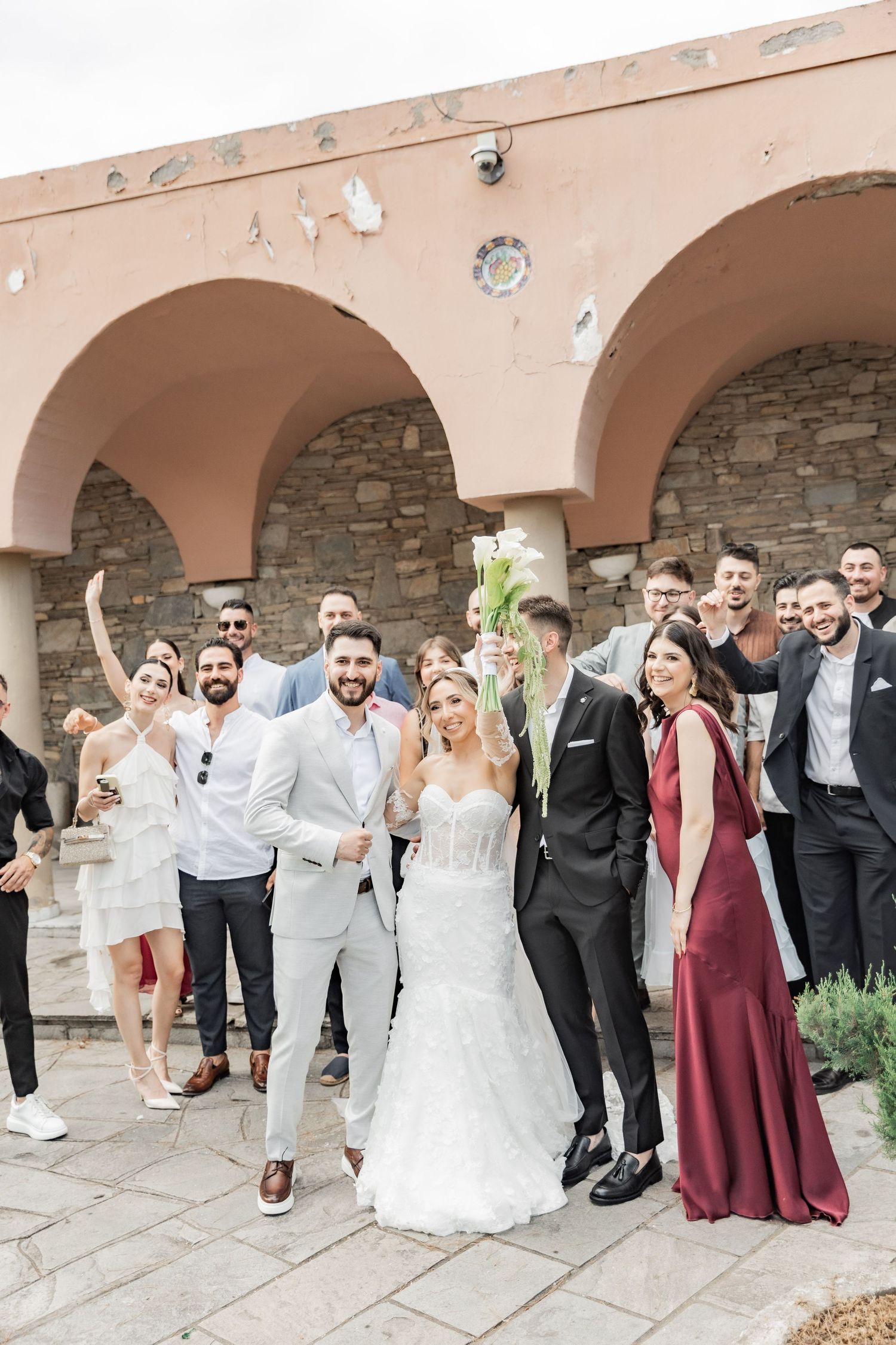 Happy wedding couple poses with guests outside a Mediterranean-style venue with arched walls.