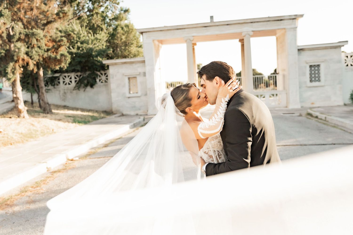A bride and groom share a romantic kiss outdoors, with the wedding veil flowing in the breeze near elegant white columns.