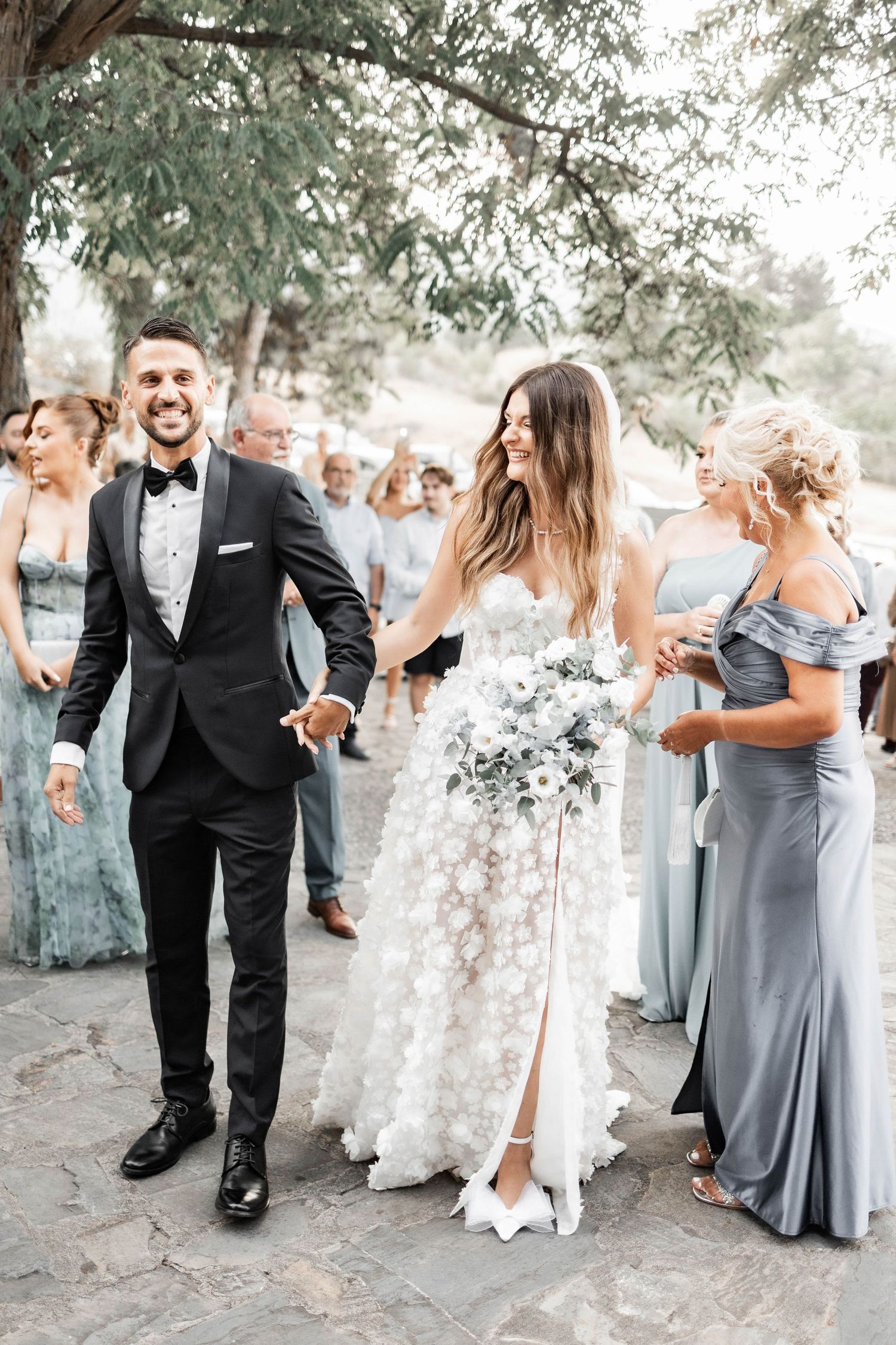 Wedding party members in gray dresses and black tuxedos celebrate outdoors under trees.